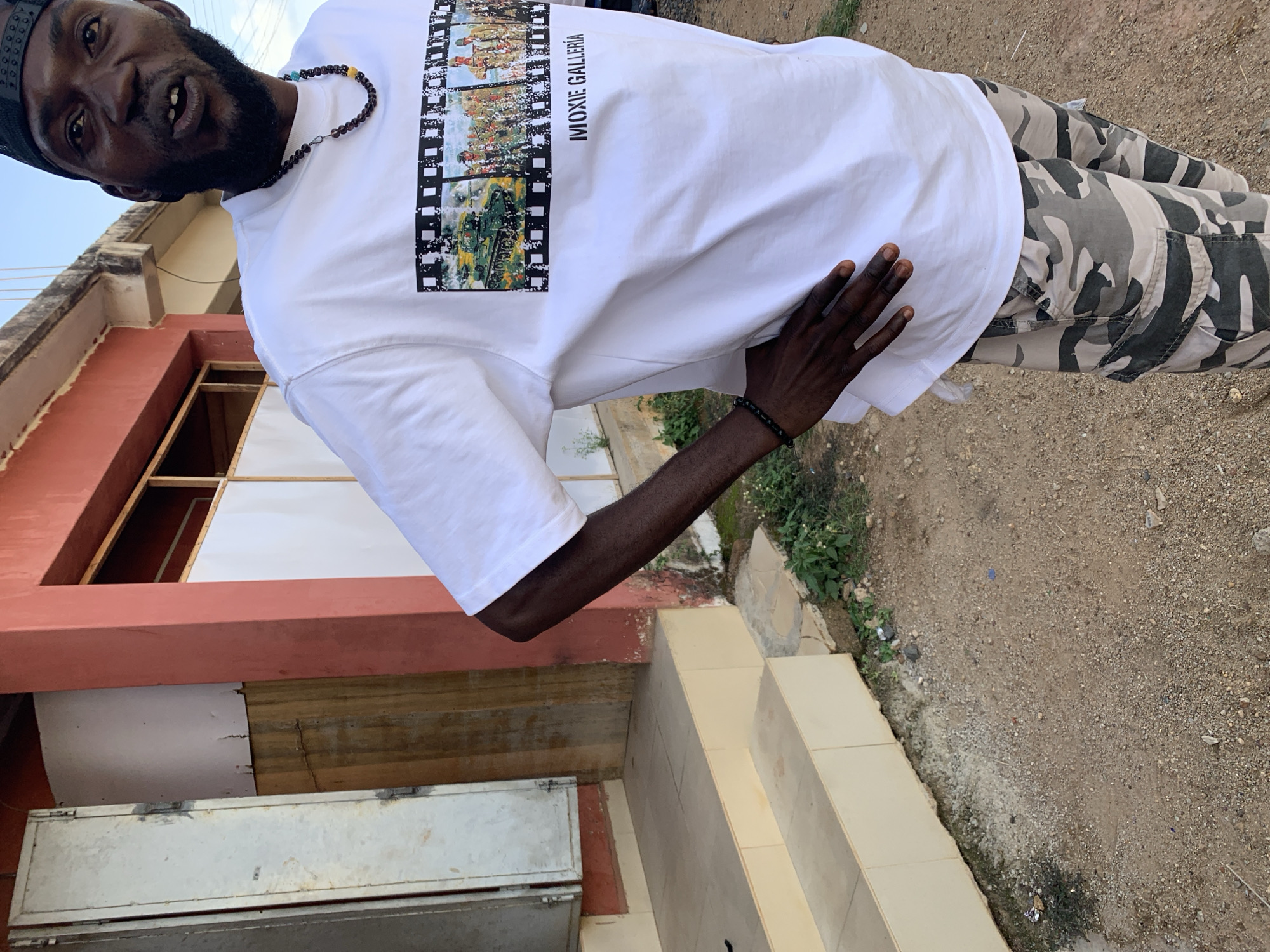 An adult Black male stands outdoors, positioned centrally in front of a building. He is wearing a black baseball cap reversed, a beaded necklace with dark, blue, and yellow elements, a white short-sleeved t-shirt featuring a film strip graphic that includes images of a green tank and soldiers, and text reading "MOXIE GALLERIA". A black beaded bracelet is visible on his left wrist. He stands with his left hand on his hip, looking slightly upward and to his right, with his mouth partially open, revealing a gap between his upper front teeth. His lower body is clad in camouflage cargo shorts.
The background consists of a building with a reddish-brown painted facade. To the left, a light-colored metal door or shutter is partially open, exposing a darker interior or wooden panel. Below this, two light beige-tiled steps lead upward. The base of the building wall shows discoloration, dirt accumulation, and sparse green vegetation. Further right along the building, a white panel with a wooden frame is present. The ground is composed of light brown and greyish dirt and gravel, interspersed with small stones and patches of green weeds. Minimal scattered debris is present on the ground. A clear, light blue sky is visible in the upper right of the frame. The scene is illuminated by natural daylight. This location is within the city of New Stadium, Nigeria.