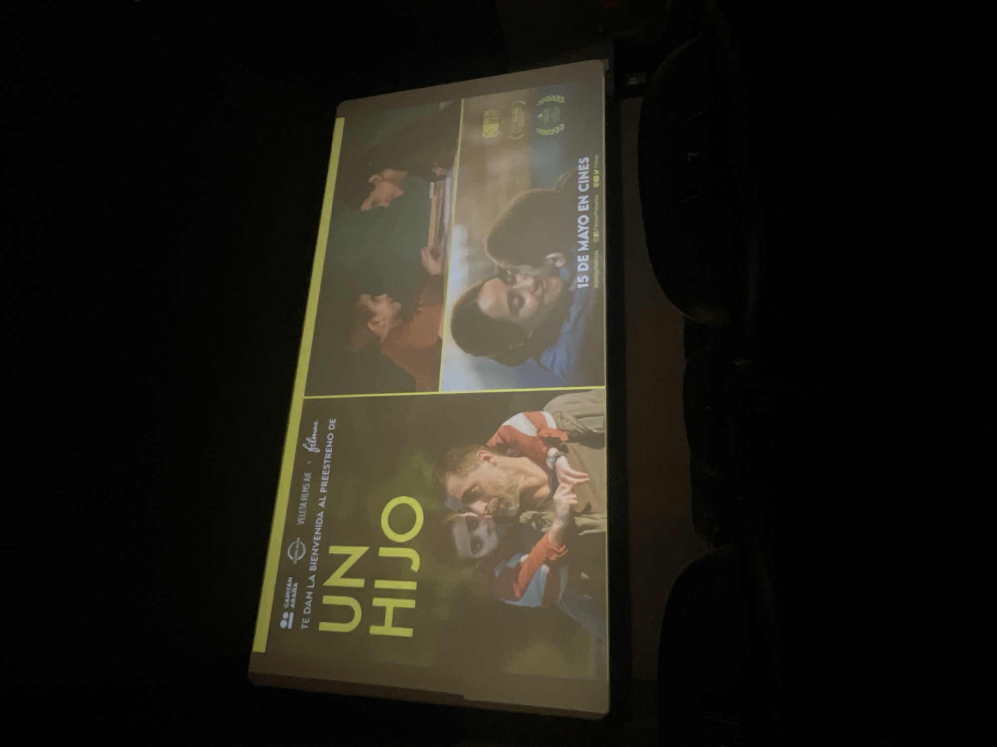 The image shows a poster or advertisement for the film "Un Hijo" on a screen, likely inside a movie theater, given the presence of theater seats in the dark background. The poster is mainly yellow and features different scenes from the film. One scene shows a man holding a child, both wearing similar striped shirts. Other scenes depict different people in what appear to be emotional moments. Text on the poster includes the film's title, "UN HIJO," the statement "TE DAN LA BIENVENIDA AL PREESTRENO DE" (which translates to "They welcome you to the premiere of"), and the information "15 DE MAYO EN CINES" (May 15th in cinemas). The logos of VELETA FILMS AIE, filmex and CAPITAN ARAÑA are also visible. The location of the image is not visible or mentioned.