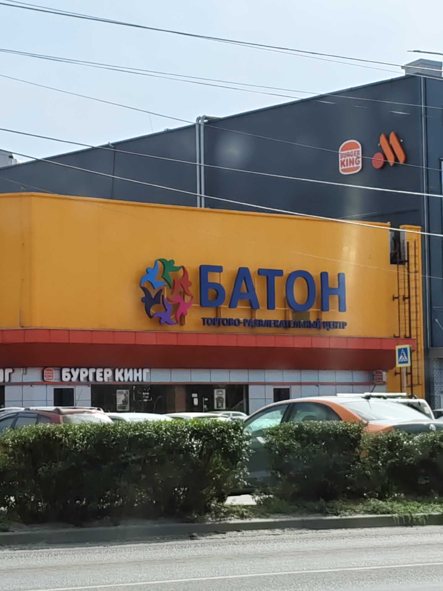 The image shows a commercial building with two prominent signs: "BURGER KING" and "БАТОН" (Baton). The "BURGER KING" sign is visible on the dark upper section of the building, with its iconic logo. Below this, a bright yellow section of the building features the large blue letters spelling "БАТОН". Beneath the "БАТОН" sign, smaller text reads "ТОРГОВО-РАЗВЛЕКАТЕЛЬНЫЙ ЦЕНТР" (Shopping and Entertainment Center). The building appears to be located in Novocherkassk, Russia. In front of the building, a row of green bushes lines a sidewalk, and cars are parked behind the bushes. The sky is visible in the background.