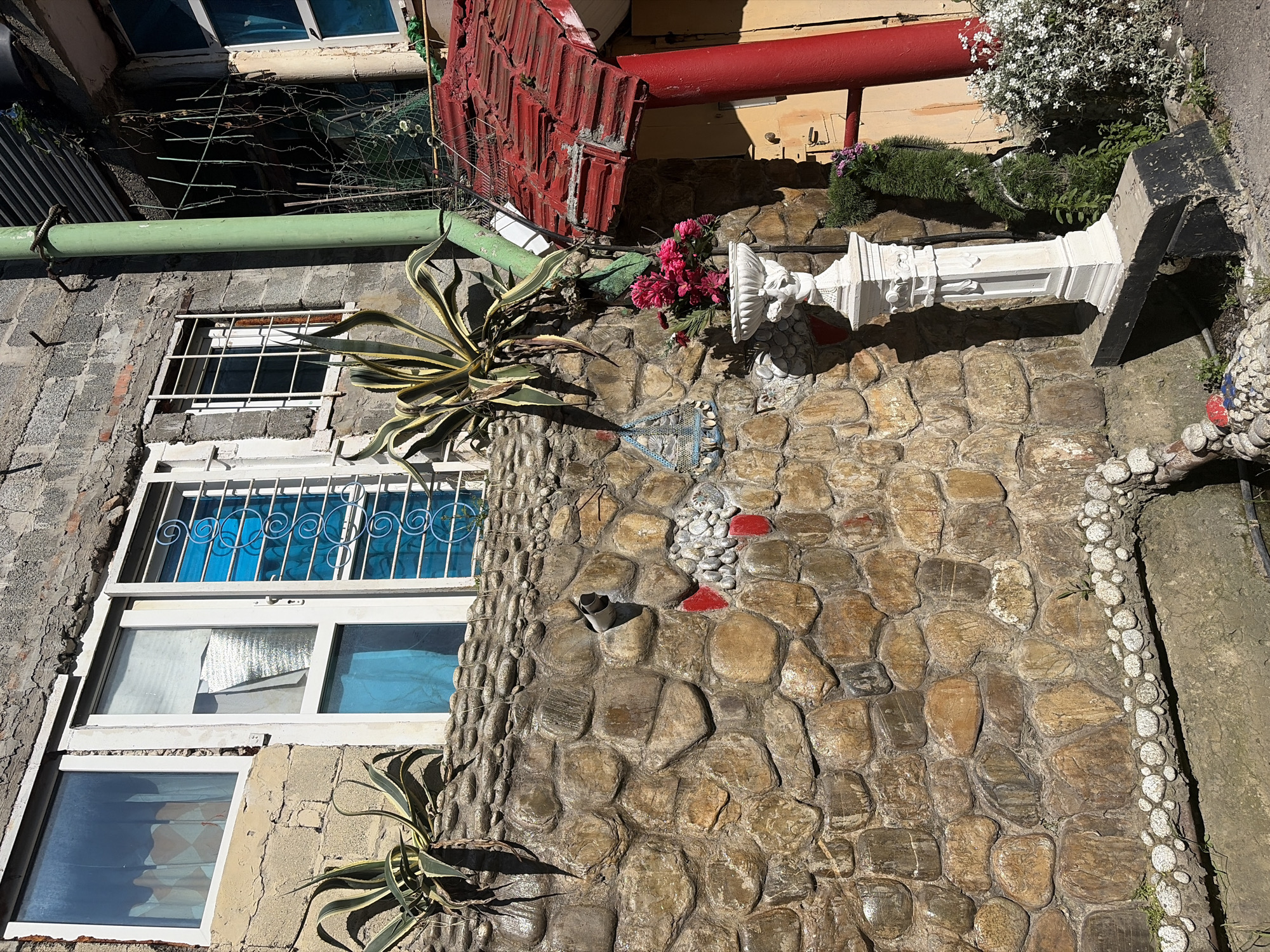 This image, captured in Sochi, Russia, presents a detailed view of a building's exterior under bright daytime sunlight. The scene is dominated by a wall featuring a diverse mix of materials and textures. The upper left section of the wall is constructed from grey concrete blocks, while a large portion of the lower and right sides is covered with a decorative facade of irregular, glossy river stones or pebbles. Some of these pebbles are painted red, and a vertical strip of smaller, rounded white pebbles forms a border on the stone wall.
Multiple windows are visible, each with distinct features. One window on the upper left has a simple metal grid, while another below it is adorned with ornate white grilles over blue-tinted glass. Further windows below show reflections of blue or white coverings from within.
The exterior is richly decorated with plants and various objects. A prominent agave plant with variegated green and yellow leaves grows from the stone wall in the mid-left. Clusters of vibrant pink flowers are placed near a white, classical-style pedestal or column, which stands on the stone wall. Another plant with small white flowers cascades from above the column on the right. Architectural elements include a horizontal green pipe running across the concrete block wall, with a segment of barbed wire or light fencing visible above it. Higher up, a thick red pipe and sections of red corrugated material, possibly part of a roof or awning, are also present. A small, gray pipe protrudes from the stone wall near the center.
No people are depicted, and no specific activity or event is occurring; the image is a static portrayal of an outdoor space. The clear, sunny weather is evident from the strong, directional sunlight and the distinct shadows cast by the various elements. No discernible text is visible anywhere in the image.