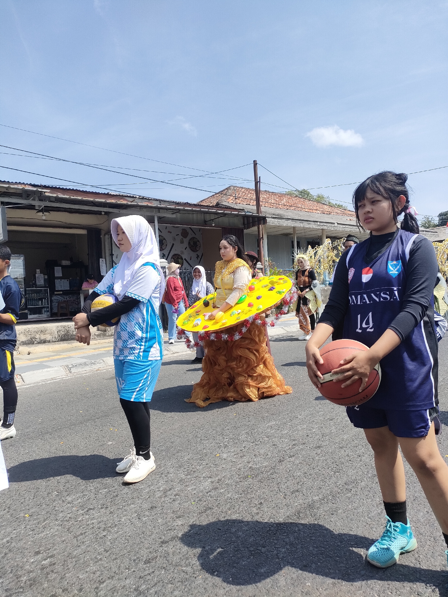 In Sungailiat, Indonesia, a group of people are participating in a parade on a sunny day. The foreground features two young women. The one on the right wears a dark blue basketball jersey with the number "14" prominently displayed and dark blue shorts, holding a basketball. The woman on the left is dressed in a white hijab and a blue and white patterned athletic outfit, holding a yellow volleyball. Behind them, a woman in a golden, elaborate dress with a large yellow circular headdress adorned with small objects and tinsel stands out. Other individuals in casual attire and traditional clothing are visible in the background, walking along a paved street lined with buildings. Power lines are strung overhead against a clear blue sky with a few scattered clouds.