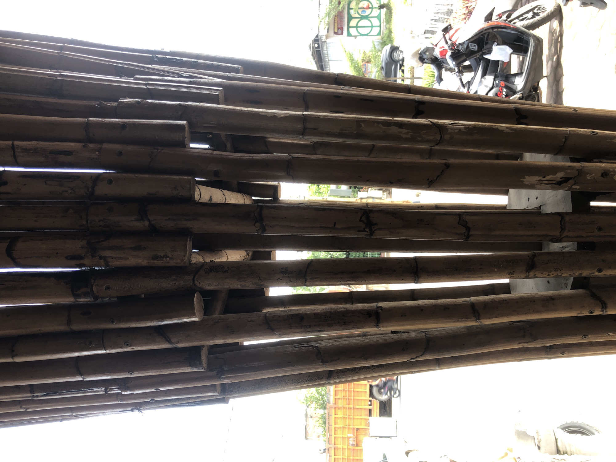 In Palohlada, Indonesia, the image presents a close-up view of a decorative structure made of vertically arranged bamboo poles. The bamboo poles, varying in thickness and exhibiting a natural weathered texture, are bundled together to form a cylindrical shape. They are spaced apart, allowing glimpses of the exterior environment through the gaps.
The outdoor scene visible through the bamboo structure includes a bright, overcast sky. In the background, there are buildings and some greenery. To the left, a large orange truck with a flatbed is parked. Further in the distance, a motorcycle is visible. To the right, another motorcycle, a scooter with red accents and the word "SCOOPY" visible, is parked on a paved surface. A dark-colored car is also present in the background on the right side of the frame. The ground appears to be a mix of paving stones and dirt. The lighting suggests it is daytime.