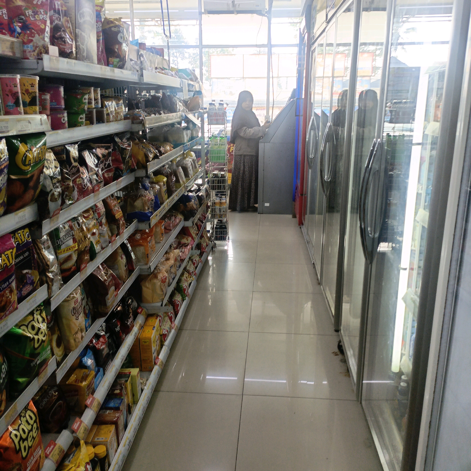 The image depicts the interior of a convenience store in Salatiga, Indonesia, specifically an aisle lined with shelves stocked with various packaged goods. On the left, shelves are filled with snacks, cereals, and other food items. On the right, glass-door refrigerators are visible, reflecting the interior and the brightly lit exterior. A woman wearing a hijab and a patterned skirt is standing at a service counter in the middle of the aisle, appearing to interact with a machine. The floor is tiled and clean, and bright fluorescent lights illuminate the space. The overall scene suggests a typical retail environment.