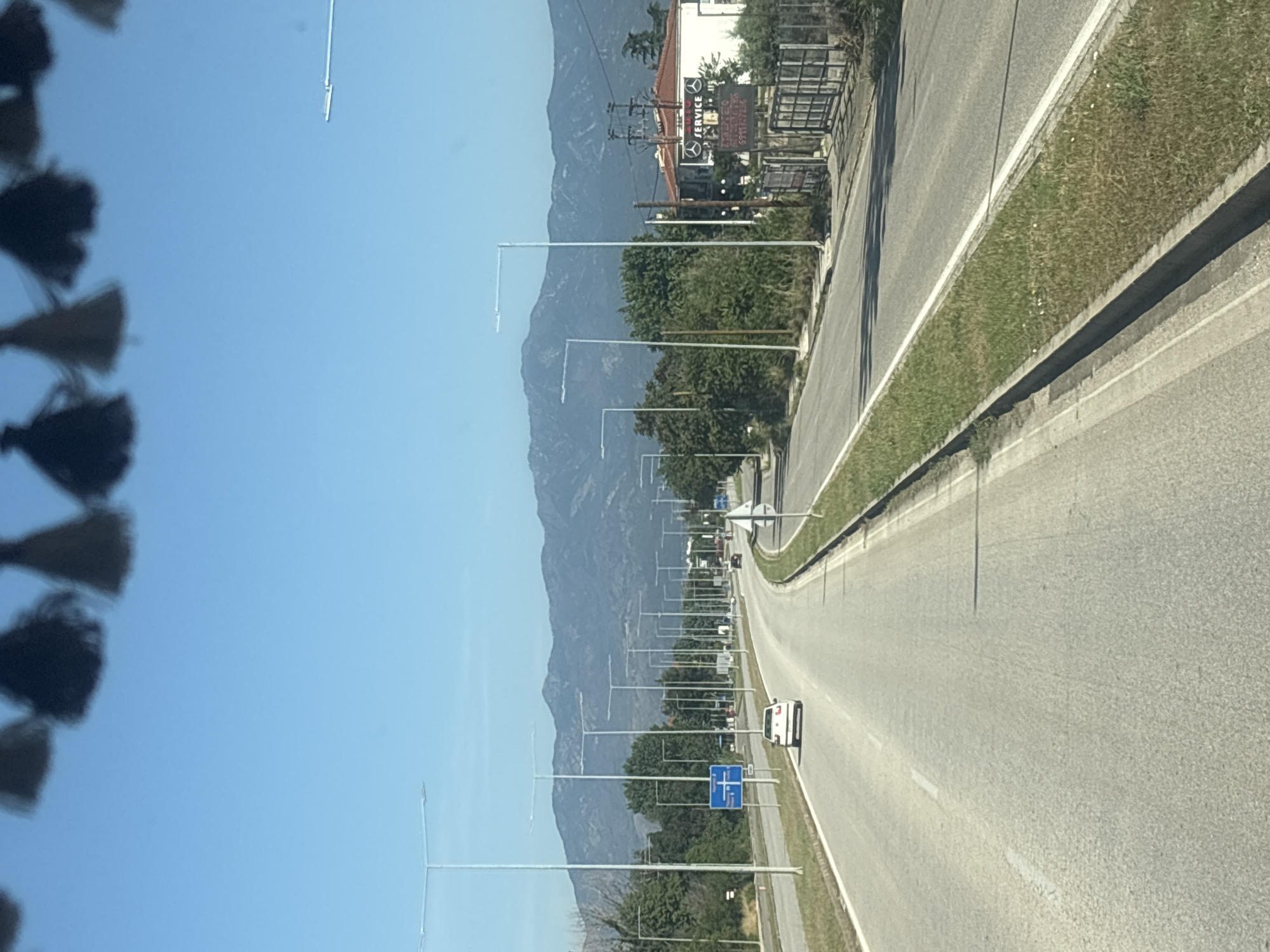 The image depicts a highway scene in the vicinity of Kikidio, Greece, on a clear, sunny day. The perspective is from inside a vehicle, looking forward along a two-lane road that curves to the right. A white car is visible in the right lane, traveling in the same direction. The roadside is lined with grass and a concrete barrier. Numerous tall, modern streetlights are positioned along both sides of the highway. In the distance, a range of mountains forms the backdrop. On the right side of the road, a building with a red roof and a sign that reads "SERVICE" is visible, along with a fenced area. The sky is a clear blue, with some dark tassels hanging from the top edge of the frame, suggesting they are attached to the vehicle's interior.