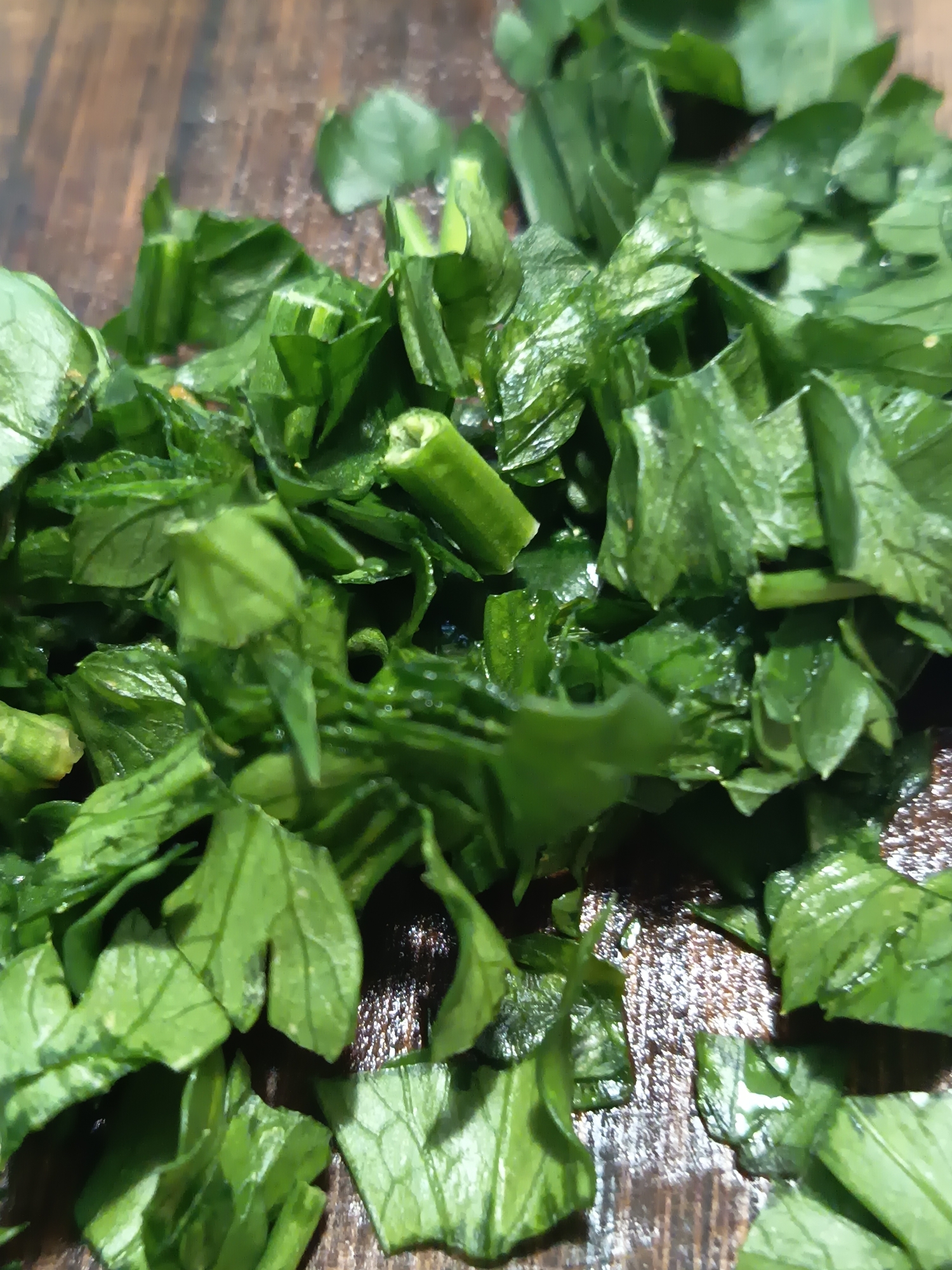 A close-up, overhead shot shows finely chopped parsley on a wooden cutting board. The parsley is bright green and appears freshly washed, with small water droplets visible on some leaves. The wooden cutting board has a visible grain pattern and appears slightly wet. The scene is lit from above, casting subtle shadows on the parsley. There are no people or other objects present. The image captures a simple culinary preparation.