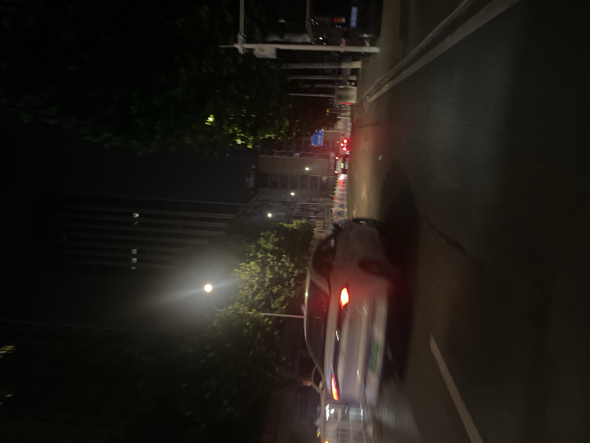 This image depicts a nighttime urban street scene within Yuzhong District, China, characterized by artificial illumination and vehicular traffic. The roadway, appearing reflective, features a prominent solid white line on the left and what appears to be double solid lines delineating a right-side lane or shoulder.
A white sedan is positioned prominently in the mid-left foreground, exhibiting significant motion blur, indicating it is moving away from the viewer. Its two red taillights are illuminated, and a blurred green rectangular element, consistent with a new energy vehicle license plate, is visible on its rear. To the immediate left of this sedan, a second white vehicle, possibly an SUV, displays illuminated headlights and also shows motion blur, partially obscured by a series of white and red barriers. Further down the road in the mid-ground, multiple vehicles are visible by their illuminated red taillights, also suggesting movement. On the far right edge of the image, a dark-colored van or SUV is partially visible, with a blue license plate partially discernible on its rear.
The environment includes tall urban buildings in the background; a multi-story structure is on the left, and a more distant, taller skyscraper-like building is in the center-right. Some building windows are illuminated. Trees with green foliage line the street, their leaves reflecting light from nearby streetlights. Street infrastructure includes utility poles, one of which supports a blue traffic sign bearing Chinese characters. Several bright streetlights are active, one intensely glowing above the foreground sedan, casting a strong light on the road and surrounding foliage. No discernible human figures are present.
