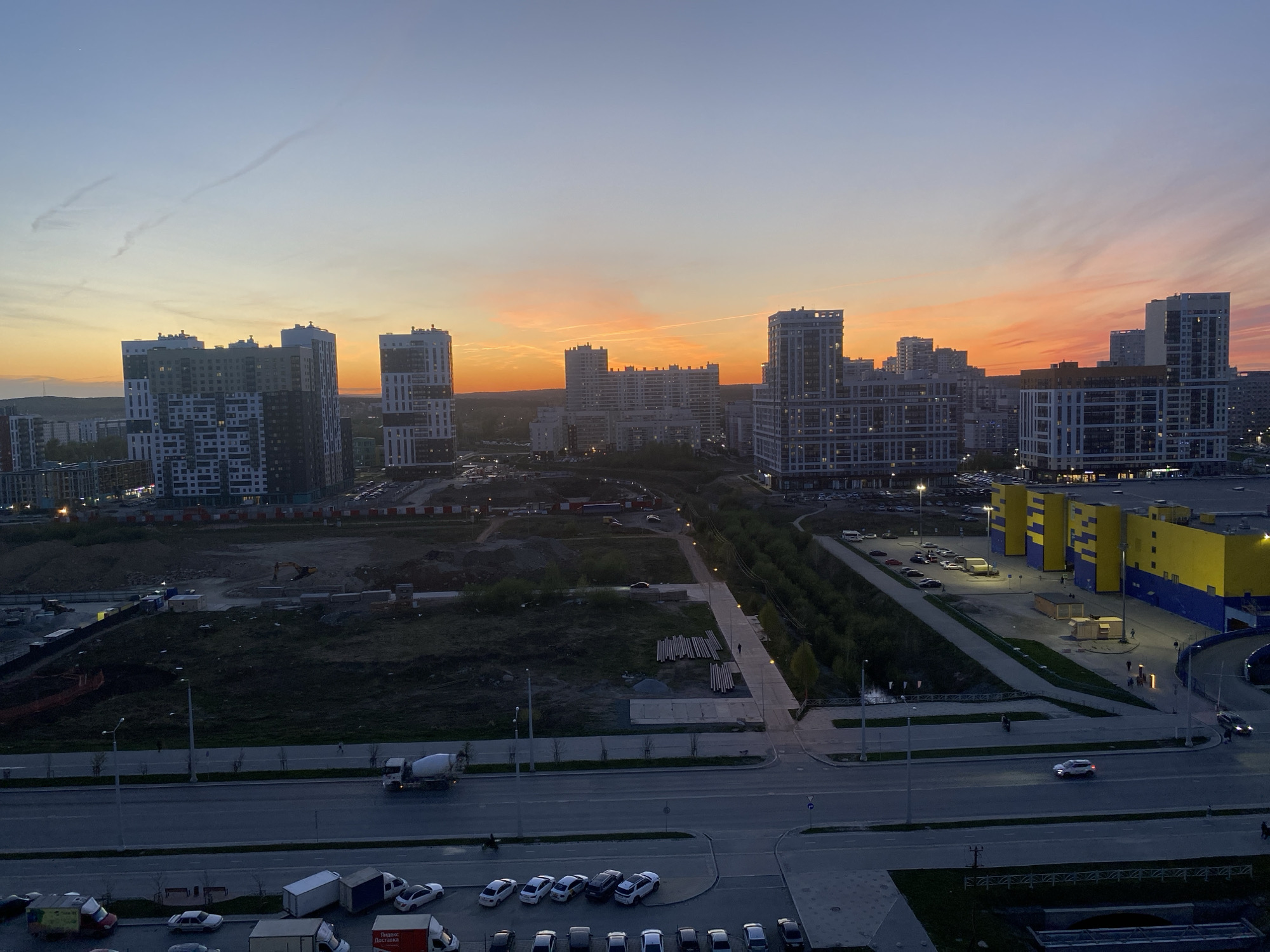 The image captures a panoramic view of Yekaterinburg, Russia, at dusk. The skyline is defined by several modern high-rise apartment buildings with mixed white and gray facades. The sunset paints the sky in hues of orange and pink above the cityscape. In the foreground, a large, vacant construction site is visible, adjacent to a road with construction vehicles. Rows of parked cars can be seen in the lower portion of the frame, suggesting a residential area. To the right, a large commercial building with a blue and yellow exterior stands out.