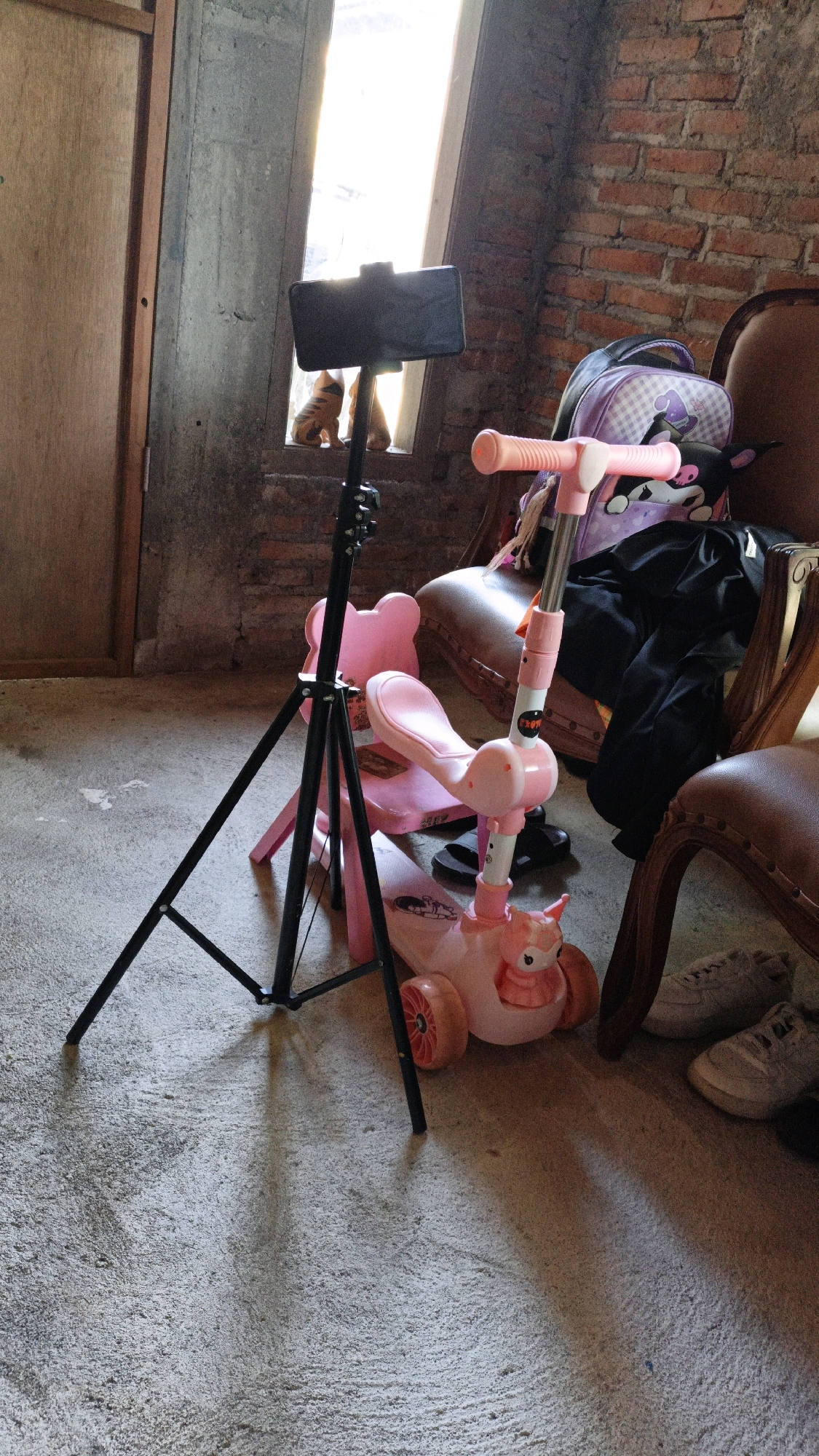 A tripod with a smartphone mounted on top stands on a concrete floor in front of a window. To the right of the tripod is a pink child's scooter with a cartoon character on the front. Behind the scooter, a purple backpack with a cartoon pattern is placed on a chair. A brick wall is visible in the background. The scene appears to be indoors, possibly in a home or a studio. The exact location in Cikal, Indonesia, is not specified in the image.