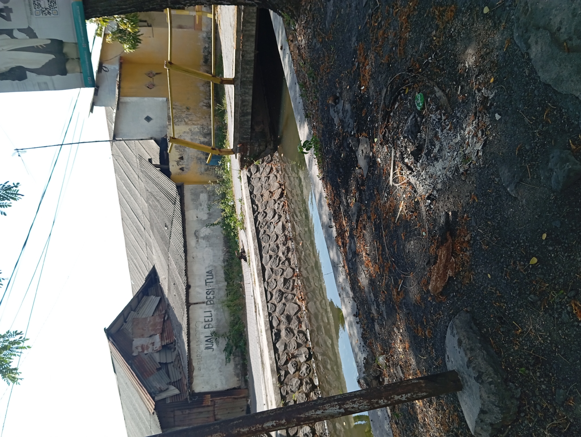 The image depicts an outdoor scene near a canal in Karangrejo, Indonesia. On a white wall, graffiti reads "JUAL BELI BESI TUA," which translates to "SELL OLD IRON." The canal runs alongside the wall, with a stone embankment on the canal's edge. A yellow bridge with metal railings spans the canal in the background, leading to a road. Buildings with corrugated metal roofs are visible behind the wall. The foreground shows a dirt and gravel area with scattered debris, including what appears to be the remnants of a fire and a green plastic bottle. A rusty metal pole is also present in the foreground, embedded in a concrete base. The overall impression is that of a somewhat neglected urban or semi-urban environment.