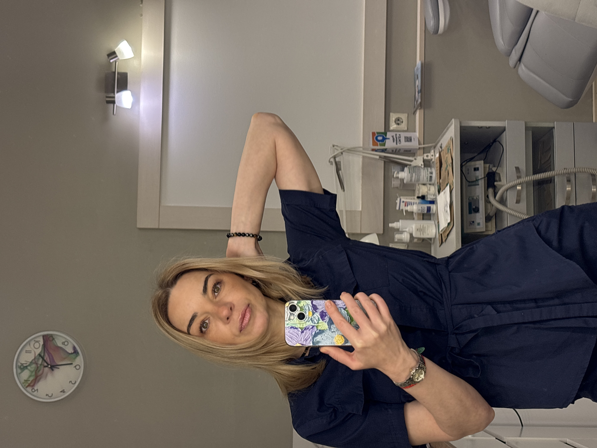 The image shows a woman taking a selfie in what appears to be a medical or dental office. She is wearing a navy blue dress, a wristwatch, and bracelets. In the background, there is a dental chair, medical equipment, a wall clock, and a wall-mounted light fixture. The location is Unknown, Unknown.