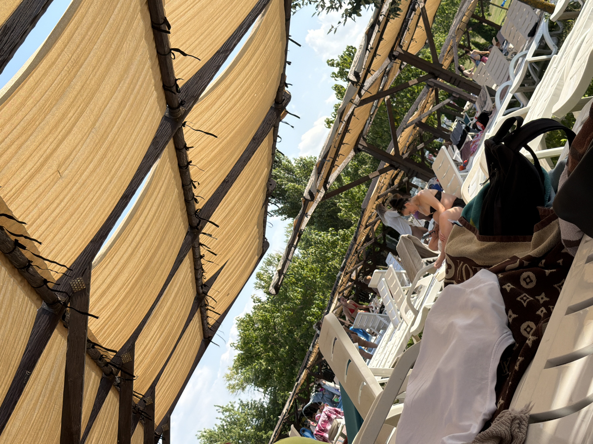This image depicts an outdoor seating area at a leisure facility, possibly a pool or resort, in the city of Sociteni, Moldova. The foreground is dominated by several rows of empty white lounge chairs, some draped with towels and bags. One chair in the lower left corner has a white t-shirt and a patterned brown towel on it. In the background, under a series of tan awnings supported by wooden structures, several people are visible. One woman in a black bikini is seated on a lounge chair. Other individuals, some in swimwear, are either walking or relaxing on chairs in the distance. Lush green trees and a bright blue sky with scattered clouds form the backdrop. The overall scene suggests a sunny day at a recreational area.