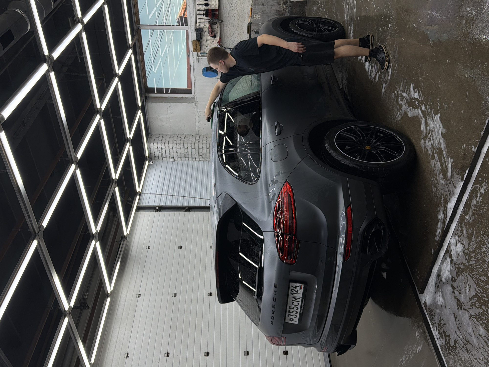 A dark grey Porsche Cayenne is parked inside a car wash in Krasnoyarsk, Russia. The car is being detailed by a young man wearing a black t-shirt and black shorts. He is using a cleaning tool to clean the roof of the car. The floor is wet and covered with soap suds. The car wash has a modern design with bright fluorescent lighting. The license plate of the car is visible and reads "P355CM 124 RUS." The overall scene suggests a professional car detailing service.
