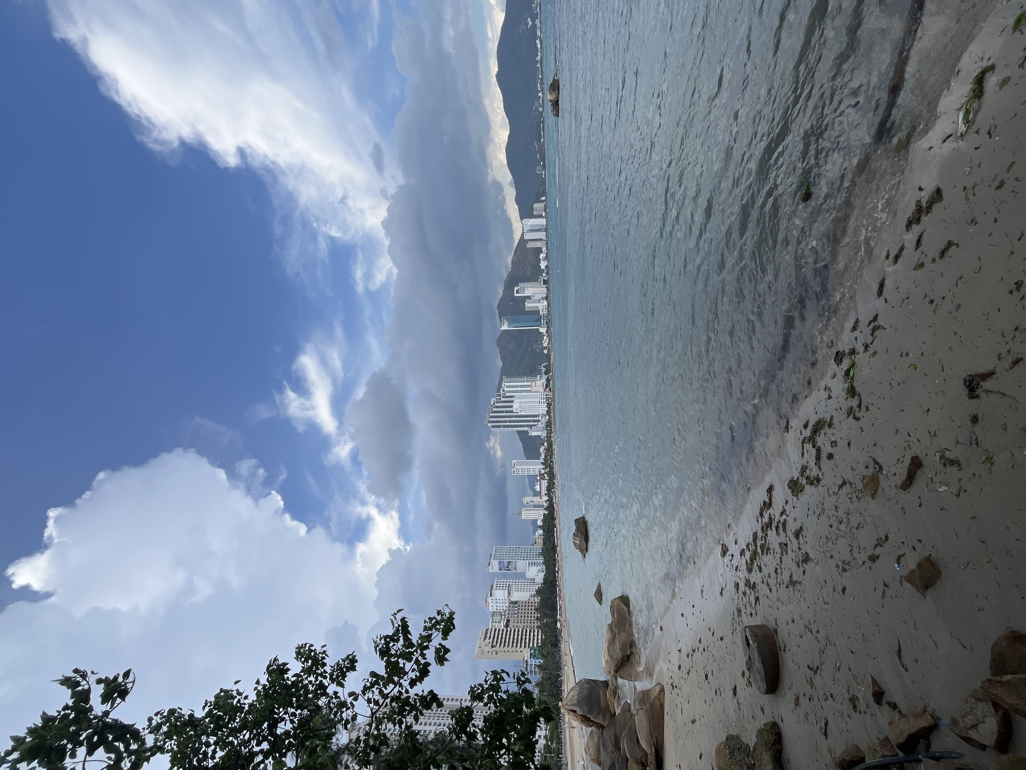 The image captures a coastal scene in Bac Nha Trang Ward, Vietnam. The foreground features a sandy beach with scattered rocks and seaweed. The water is murky, possibly due to the tide or recent weather. A line of high-rise buildings stretches along the coastline, indicating a developed urban area. Behind the buildings, a mountain range is partially obscured by clouds. The sky is a mix of blue and grey, with puffy white clouds on one side and darker, stormy clouds on the other. A few trees and buildings are visible in the bottom left corner, framing the scene.