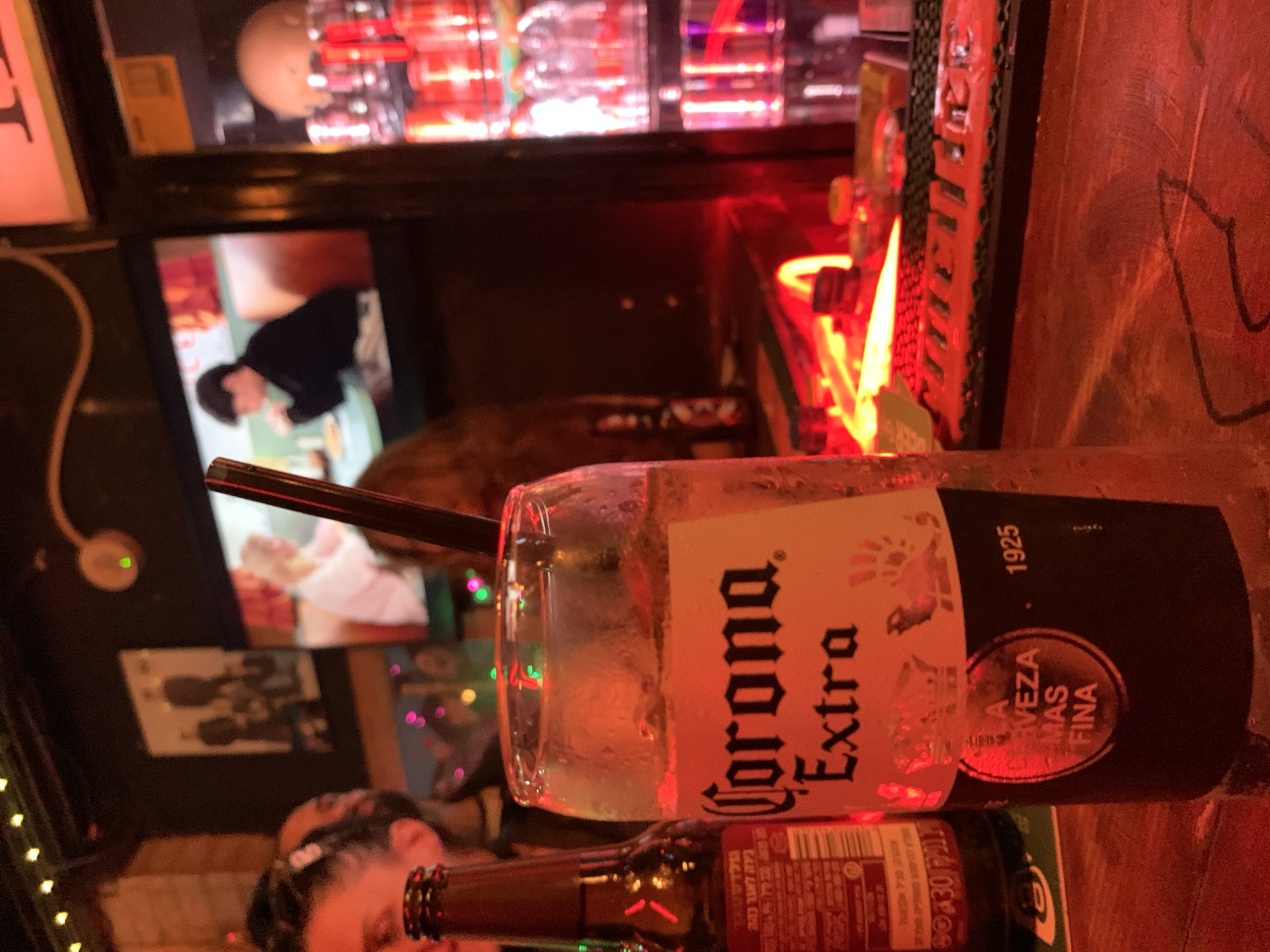 The scene is an indoor bar, likely in Tel Aviv, Israel, given the presence of a Corona Extra beer bottle and drink. The bar is dimly lit with red lighting. In the foreground, a partially full glass of Corona Extra sits next to an open Corona Extra beer bottle. A black straw is in the glass. In the background, there is a large flat screen showing what appears to be a person sitting at a table. Above the screen is a shelf with assorted bottles and glassware, also illuminated by red light. Further in the background and partially obscured, is a person or people at the bar, one with braided hair. There are lights on the left side. The bar surface appears to be wooden with some graffiti.