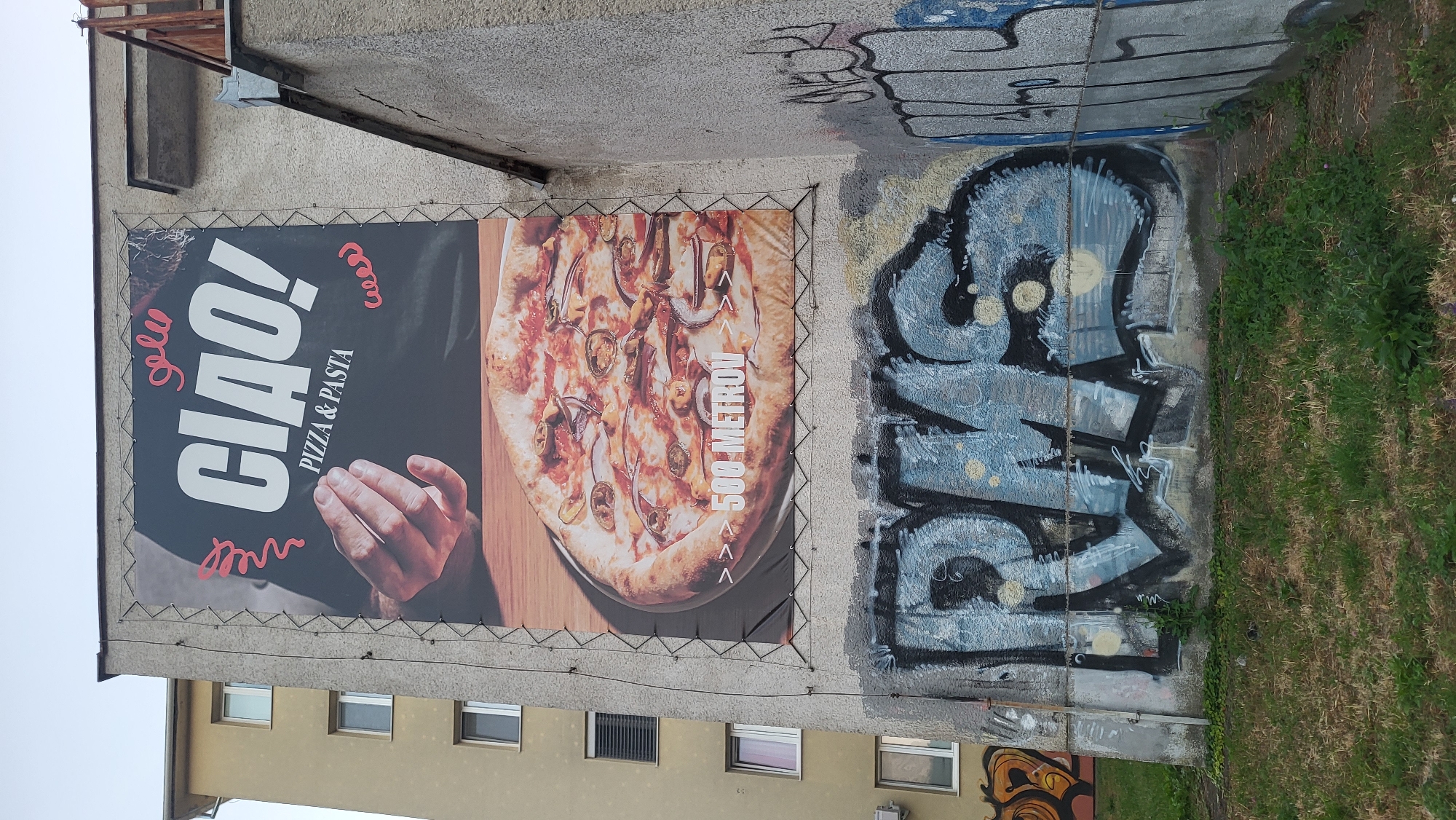 This is an eye-level, full, medium outdoor shot of a large advertisement and graffiti on the side of a building. The advertisement, which is for a pizza and pasta restaurant called "CIAO!", features a close-up of a pizza and the text "500 METROU" (meaning 500 meters in Slovak). The advertisement is mounted on a rough, grey concrete wall of a multi-story building. The building appears to be an apartment building, with several windows visible on the left side of the frame. Below the advertisement, the wall is covered in graffiti, with the most prominent tag being "RMS" in large, blue and black spray paint. To the left of the "RMS" tag, there is another smaller graffiti tag with orange and black colors. The ground in front of the building is covered in patchy green grass and some dry patches. The sky above is overcast and a pale blue. The scene is likely in an urban environment, possibly a residential area in Prešov, Slovakia, given the Slovak text on the advertisement. There are no people visible in the image.