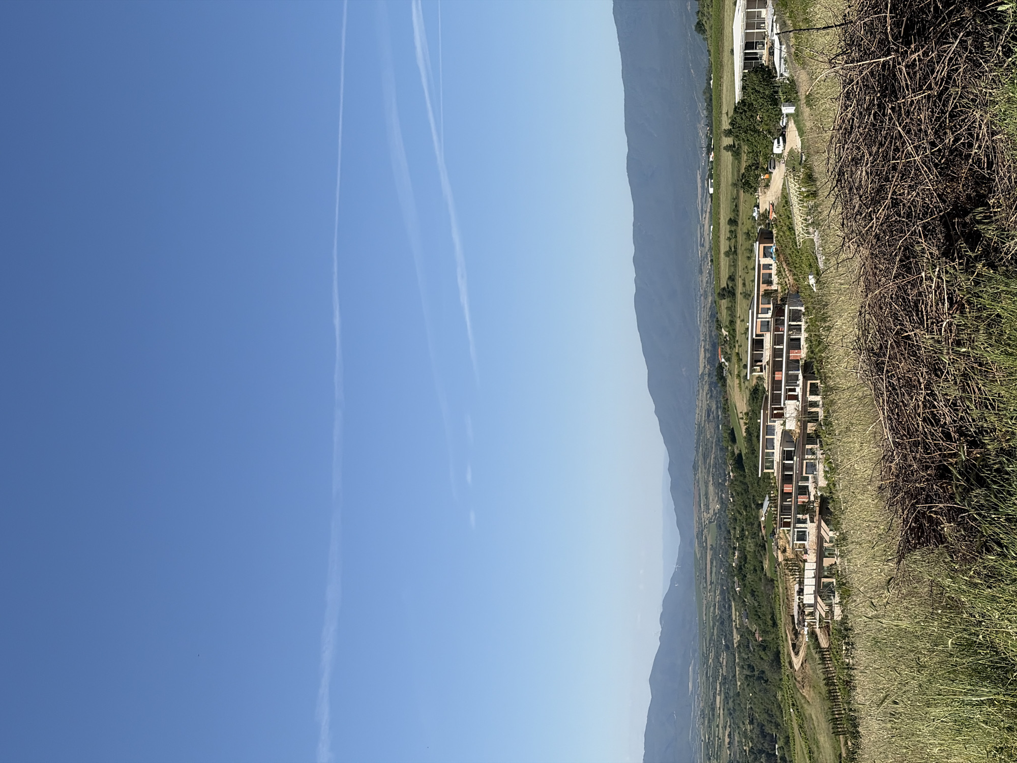 The image shows a mountainous landscape. The sky is clear blue with contrails. A range of mountains is visible in the middle distance. A cluster of modern buildings is situated in the valley. In the foreground, there's dry grass and a pile of branches.
Location: Unknown, Unknown.