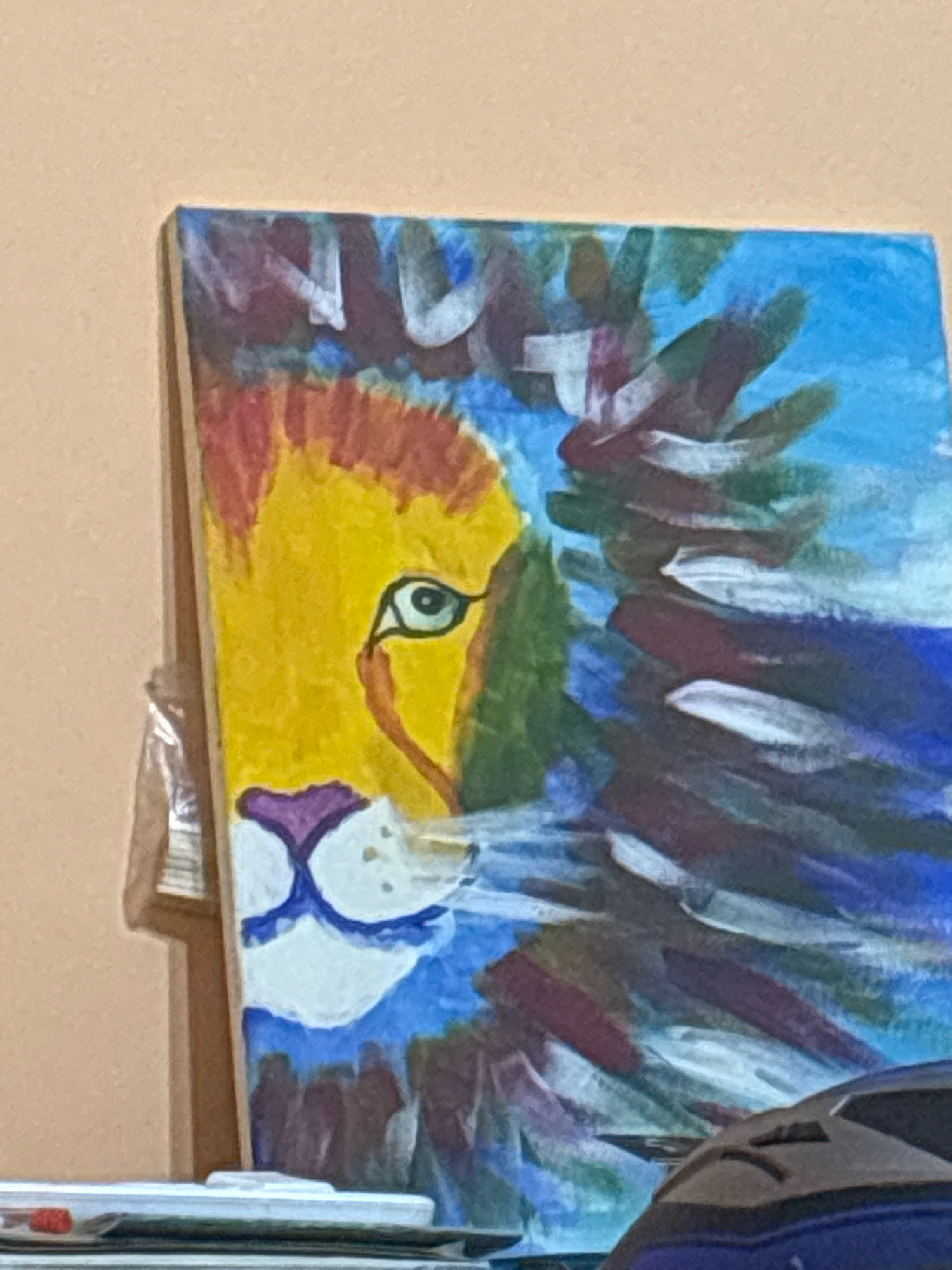 The image shows a close-up of a painting of a lion's face. The lion has a yellow face with a green eye and a purple nose. The mane of the lion is depicted with strokes of red, blue, and purple. The painting is abstract and colorful. The painting is leaning against a light brown wall. In the foreground, a portion of a dark blue object is visible, possibly a bag or backpack. There are also some white objects that appear to be part of a shelf or desk in the foreground. The image is taken from a slight angle. No specific location in Krichim, Bulgaria can be identified from this image.