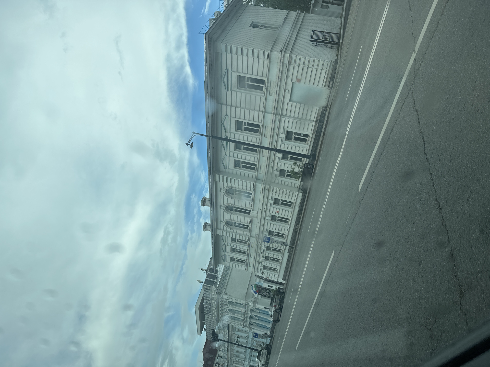 The image shows a street scene in Ufa, Russia, on a cloudy day. The primary focus is on a row of white, historic buildings lining the right side of the street. The architecture appears to be classical, with arched windows and decorative cornices. Several cars are parked along the street in front of the buildings, and there is a pedestrian walking in front of one of the structures. A street lamp stands prominently in front of one of the buildings. The road in the foreground is asphalt with white lane markings. The sky is filled with a mix of clouds and patches of blue sky. The overall impression is of a well-maintained urban environment with older architectural heritage.