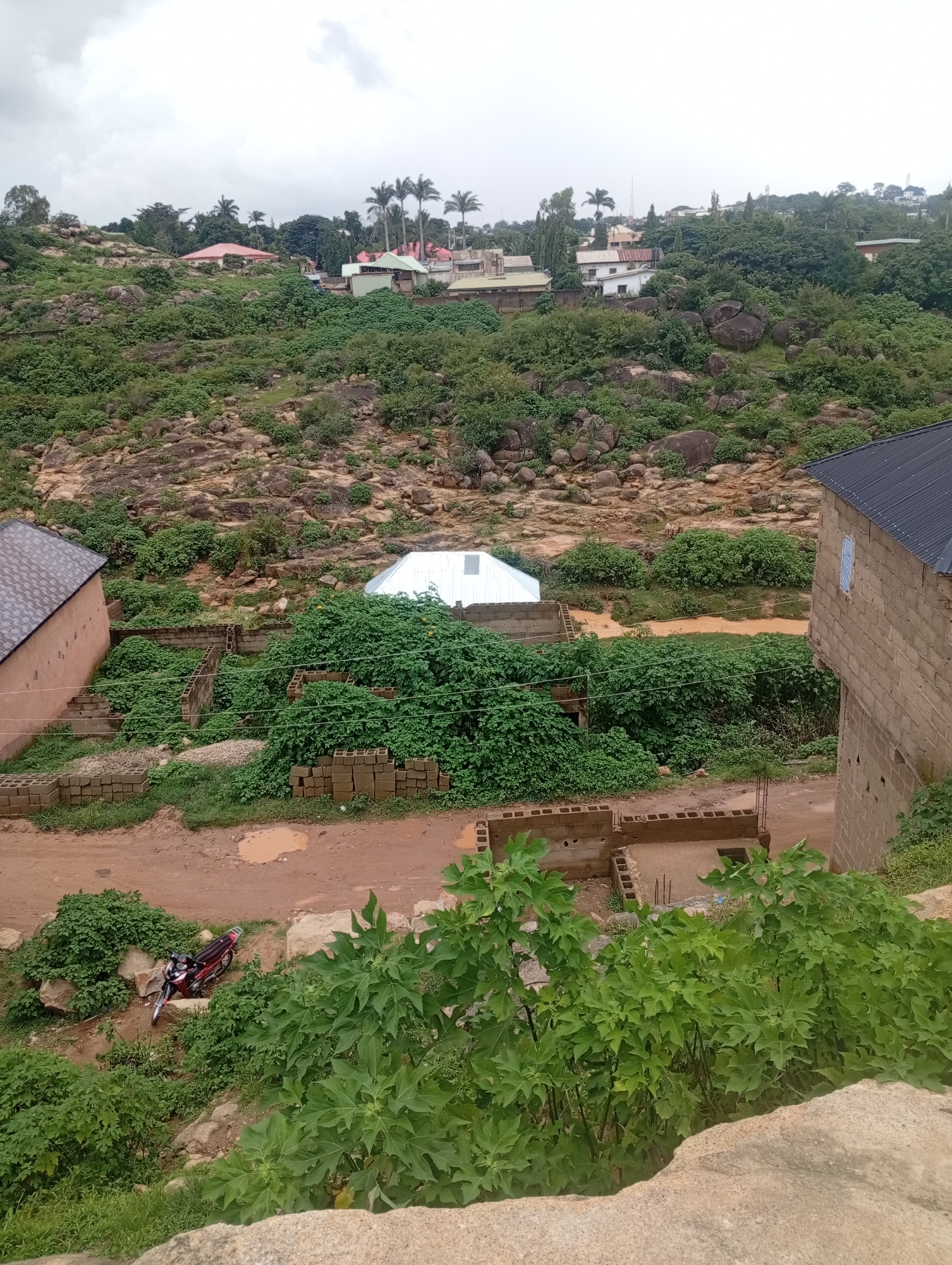 The image depicts a rocky, overgrown landscape with scattered buildings in the city of Jos, Nigeria. In the foreground, a red motorcycle is parked on a dirt path next to a clump of green bushes. Further into the scene, there are unfinished brick structures surrounded by dense vegetation, indicating ongoing construction. A white, pitched-roof building is visible in the middle ground, and beyond that, a cluster of residential buildings with red and green roofs sit atop a rocky hill. Palm trees and other trees dot the landscape, and the sky is overcast. The overall impression is of a somewhat undeveloped or rural area within the urban setting of Jos, characterized by its hilly terrain and abundant greenery.