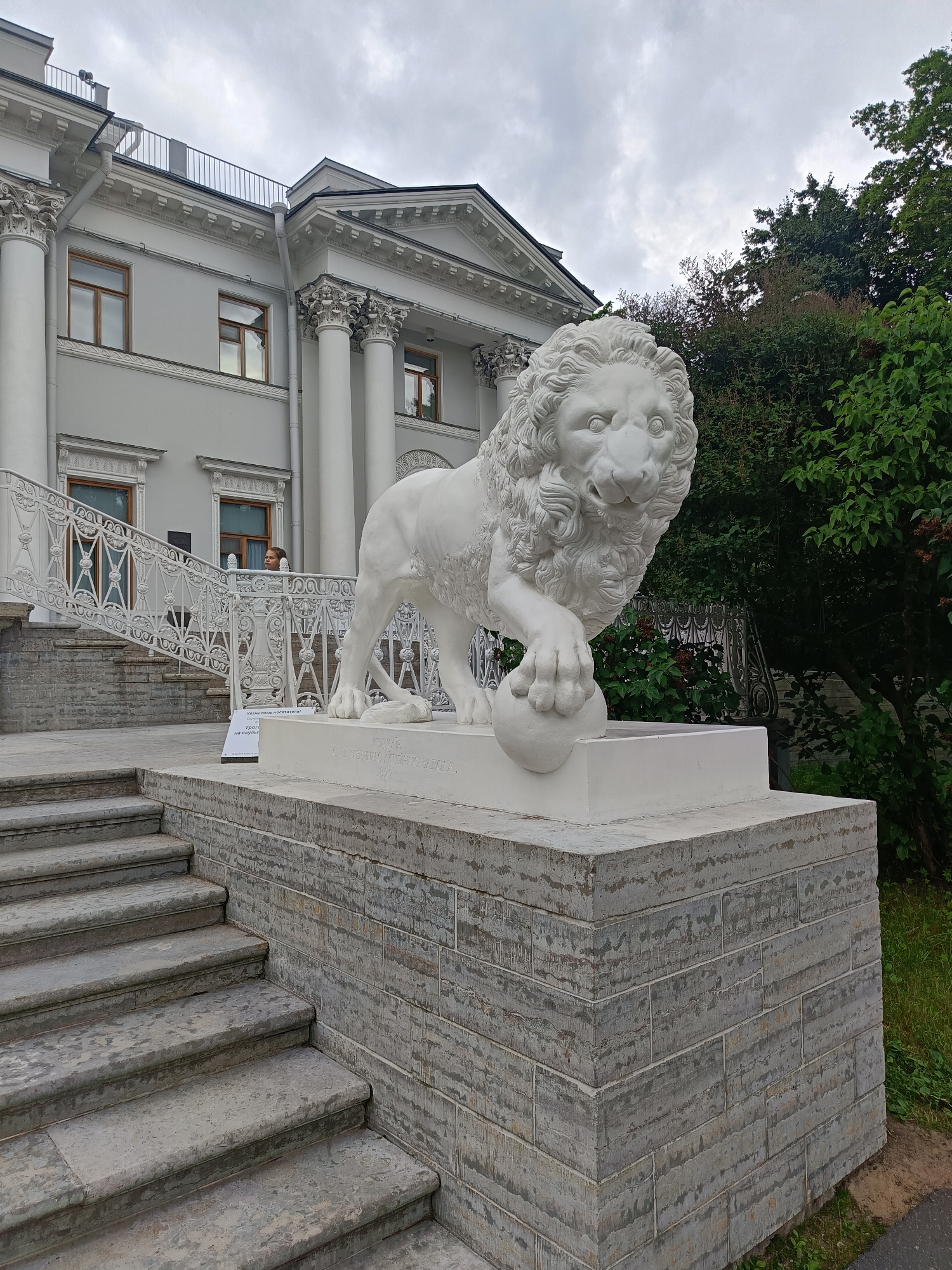 A white sculpture of a lion with a ball at its paw stands on a pedestal in front of a classical building in Saint Petersburg, Russia. The building features large columns, ornate detailing, and large windows. A set of stone stairs leads up to a white, decorative railing. A person is visible behind the railing. The lion sculpture is detailed, with a mane, expressive eyes, and muscular legs. The surrounding environment includes green trees and a cloudy sky. A sign is placed in front of the lion sculpture on the pedestal.