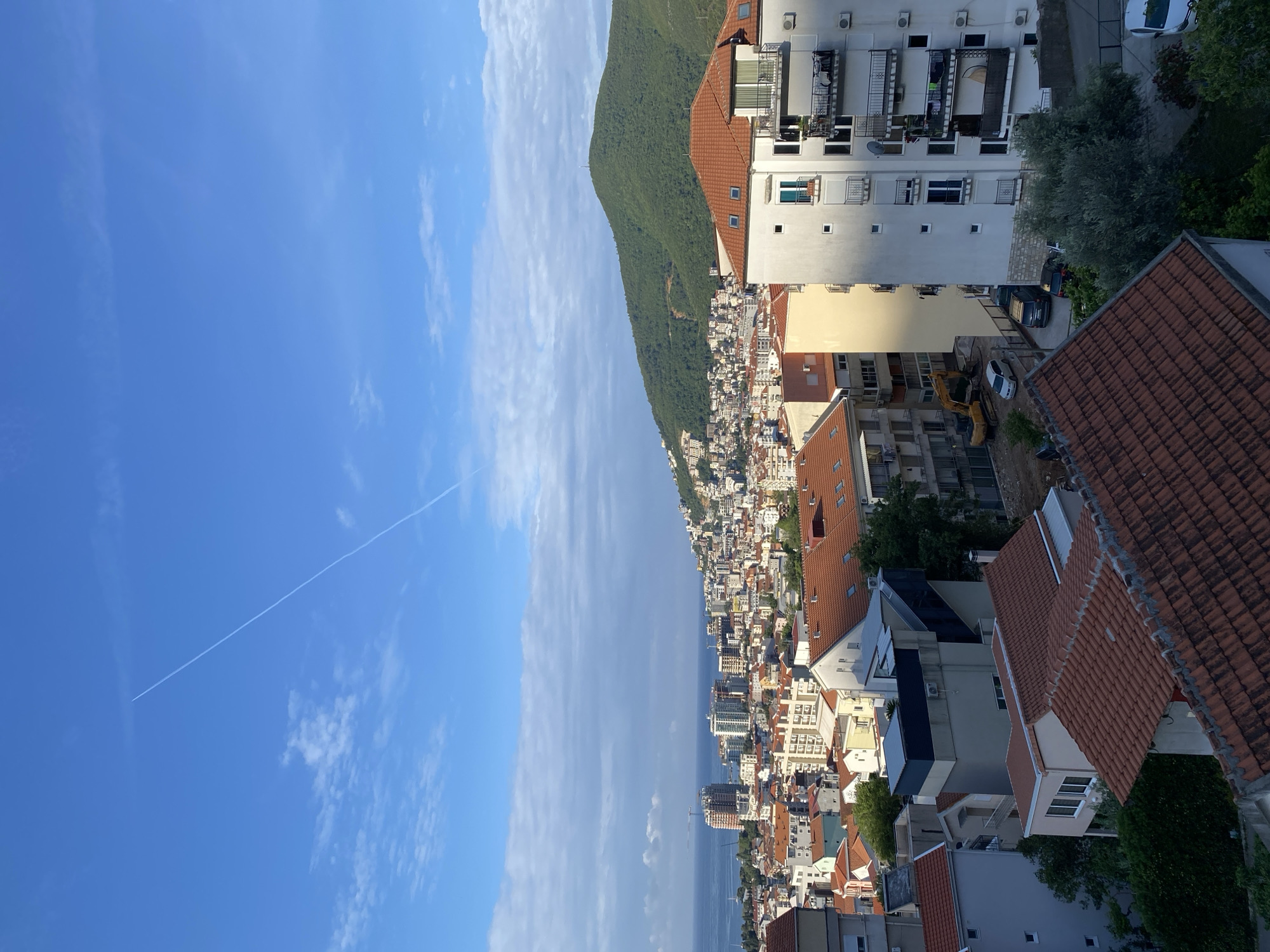 The image shows a coastal city in Budva, Montenegro, nestled between a mountain and the Adriatic Sea. The sky is blue with a few clouds, and a contrail is visible. The city is densely packed with buildings, mostly with red-tiled roofs. There is a hill covered in lush greenery next to the city. The coast is lined with buildings and infrastructure. In the foreground, rooftops and vegetation are visible.