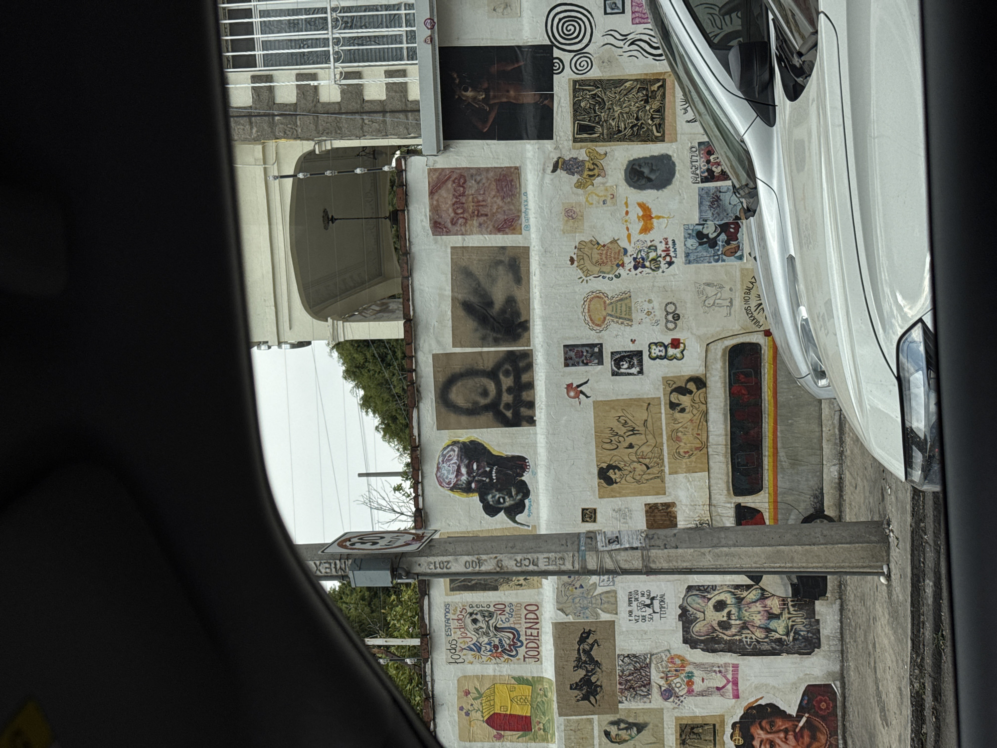 A view from inside a car shows a stucco wall heavily decorated with posters, drawings, and graffiti. The wall appears to be part of a building facade next to a street. Some posters depict faces, figures, and abstract designs. A white car is parked near the wall, partially obstructing the view of some posters. A utility pole with a speed limit sign is visible, marked with "MEX" and "CFE DCR 9 400 2013." The speed limit is "30". Several phrases and words are discernible, including "ESTAMOS TODOS JODIDOS", "JODIENDO", "SOMOS", "MAJITO", "PULKAZOS NO BALAZ," "Y POR PRIMERA DESIO QUE ESTO NO SEA ALGO TEMPORAL," "Silence Honey." The image suggests a street art scene likely located in Puebla, Mexico.