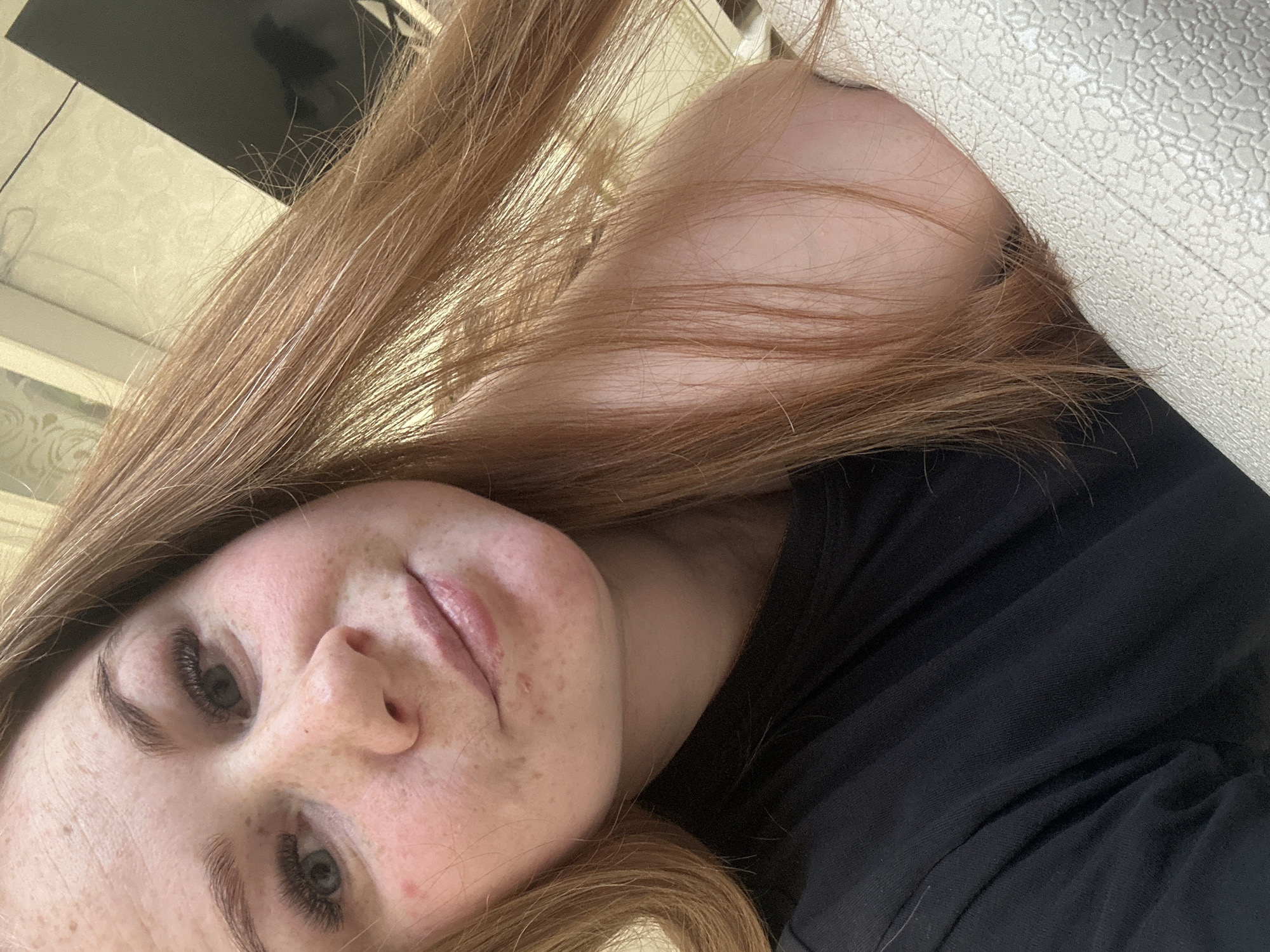 A close-up, angled shot of a fair-skinned woman with red hair. She is likely lying down, as her face is tilted. She has freckles and appears to be wearing false eyelashes. She is wearing a black top. The background includes a textured white surface, possibly a wall or headboard, and a glimpse of a window. The location is not specified. The city of Chaikovsky, Russia, cannot be confirmed as the image does not contain any identifying features of this location.