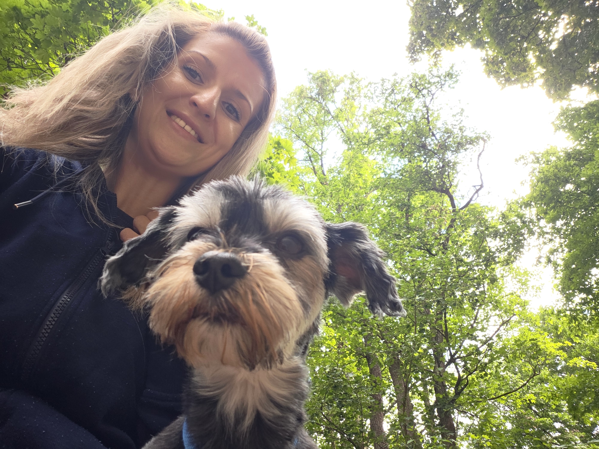 This is a low-angle, outdoor selfie taken from below a woman and a dog. The woman, who has blonde hair and is wearing a dark blue zippered jacket, is smiling and looking towards the camera. She is holding a small, dark-colored dog with tan markings in front of her. The dog's face is blurry and positioned in the foreground. The background consists of lush green trees with sunlight filtering through the leaves, suggesting a park or forest setting. The sky is bright and overcast, indicating daytime. There are no discernable landmarks to specify a precise location within Munich, Germany.