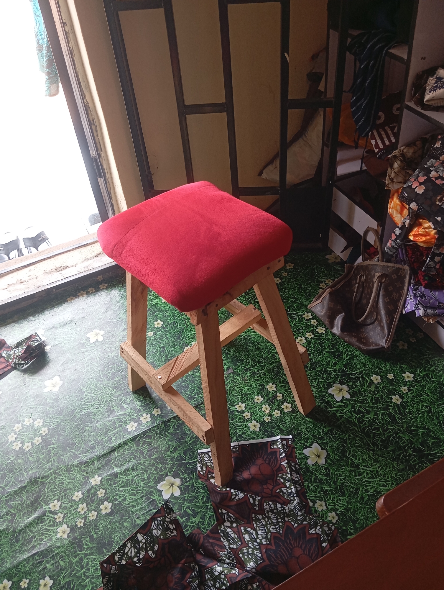 In an indoor setting, a red cushioned stool with a wooden frame stands on a floor covered with a green grass and flower patterned sheet. To the right of the stool, a brown designer handbag rests on the floor. Behind the stool, a shelving unit is visible, containing various clothing items and bags. The scene is illuminated by natural light streaming in from a window to the left, which has metal bars across it. The background appears to be a wall with a wooden door frame. The overall impression is that of a storage or dressing area.