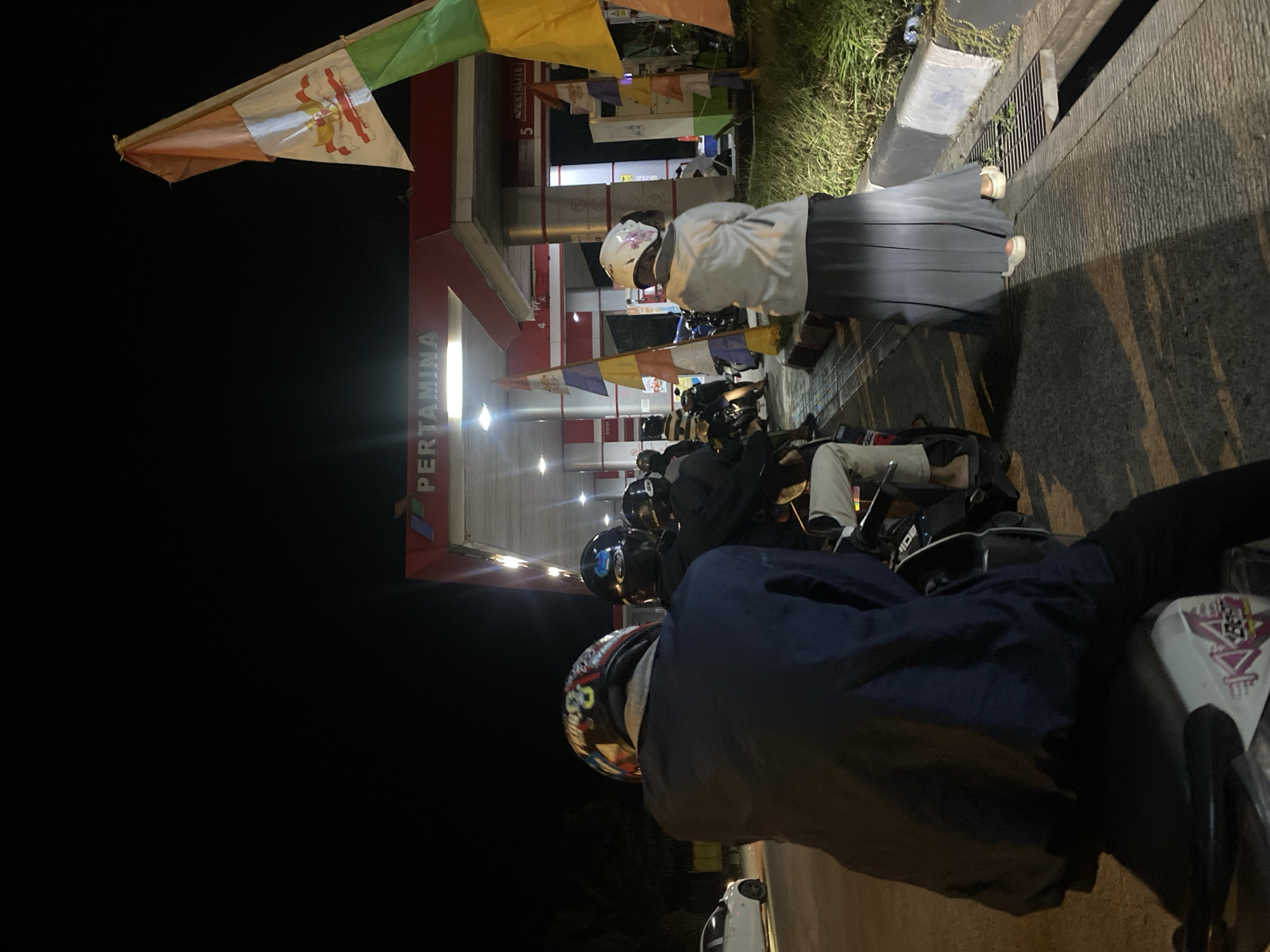 At night, an image depicts a Pertamina gas station in Gambut Barat, Indonesia. Several people on motorcycles are waiting in line. The foreground features a person wearing a dark blue jacket and a colorful helmet, seated on a motorcycle. To their right, another person in a white hijab and a long gray skirt is also visible. The gas station is illuminated by overhead lights, and flags are displayed on poles. In the background, other motorcycles and people are present, and a white car is parked on the side of the road. The environment appears to be a typical urban street scene at night.