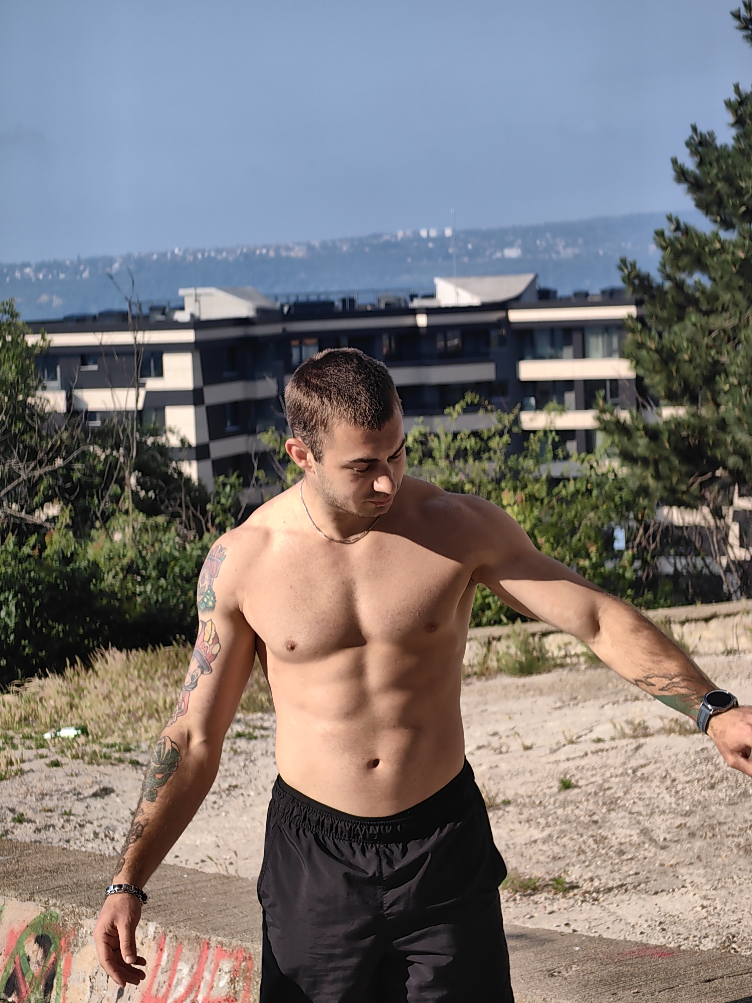 A shirtless man with short brown hair, defined abdominal muscles, and tattoos on his left arm stands outdoors. He is wearing black shorts and a silver necklace. A silver bracelet is on his left wrist, and a dark watch is on his right wrist. He is looking down and to his left, with his left arm bent at the elbow and his right arm extended out of frame. The background features a modern building with dark and light gray facades and white accents, surrounded by green trees and bushes. In the distance, a hazy blue sky meets a distant cityscape and a body of water. The foreground shows a concrete surface with graffiti and dry grass. The scene is likely located in Varna, Bulgaria, given the characteristic architecture and landscape.