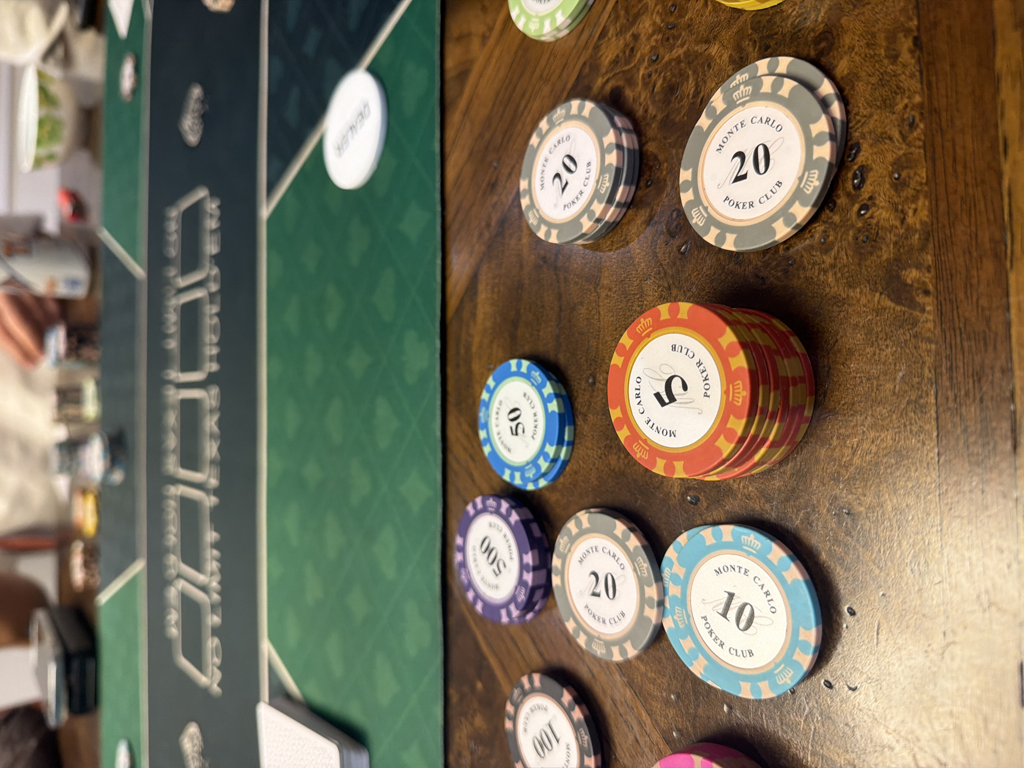The image shows a close-up of a poker table and chips on a wooden surface. The poker table is green with "NO LIMIT TEXAS HOLD'EM" printed in white letters. Several stacks of poker chips are visible, with denominations of 5, 10, 20, 50, 100, and 500. The chips are branded "MONTE CARLO POKER CLUB". A white "DEALER" chip is also visible on the green felt. In the background, blurred figures of people can be seen, along with some drinks and a bowl. The scene appears to be set indoors, likely in a home or casino. There is no indication of the image being located in Xuân Trường - Đà Lạt, Vietnam.