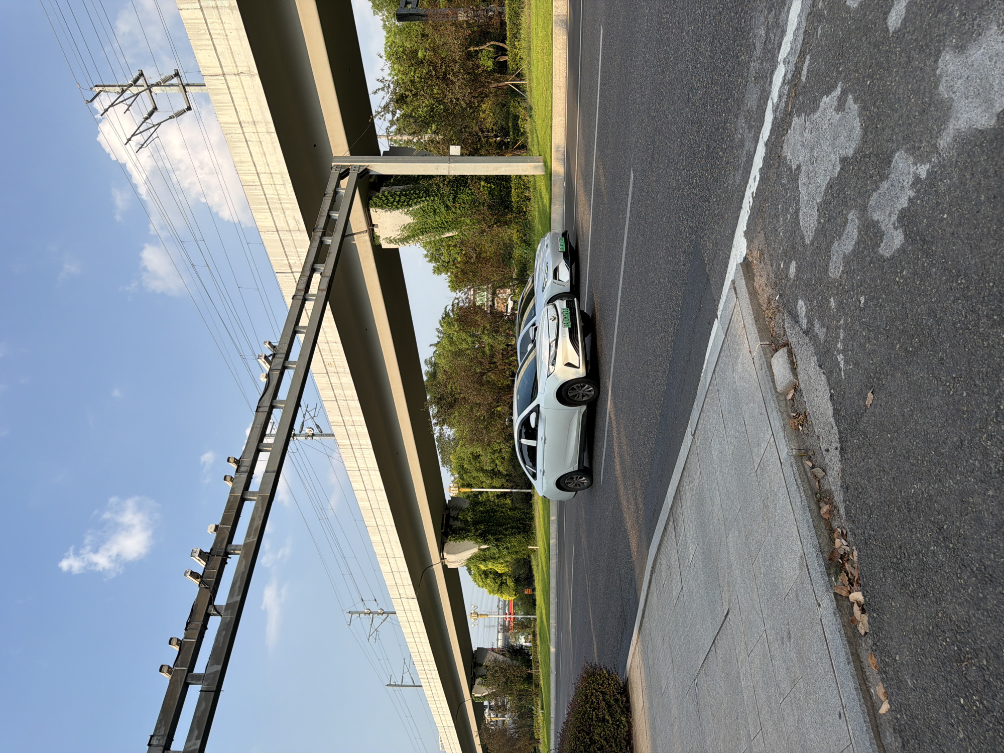 In the city of Jiangjin, China, two white electric cars are driving on an asphalt road under a large elevated structure, likely an overpass or elevated railway. The sky is blue with a few scattered white clouds. The elevated structure is supported by concrete pillars, one of which is covered in green ivy. Overhead, there are electrical wires and poles. On the left side of the road, there is a sidewalk made of large gray tiles, with some dry leaves scattered near the curb. The road has clear white lane markings. The scene appears to be a typical urban or suburban environment with infrastructure for transportation and power.