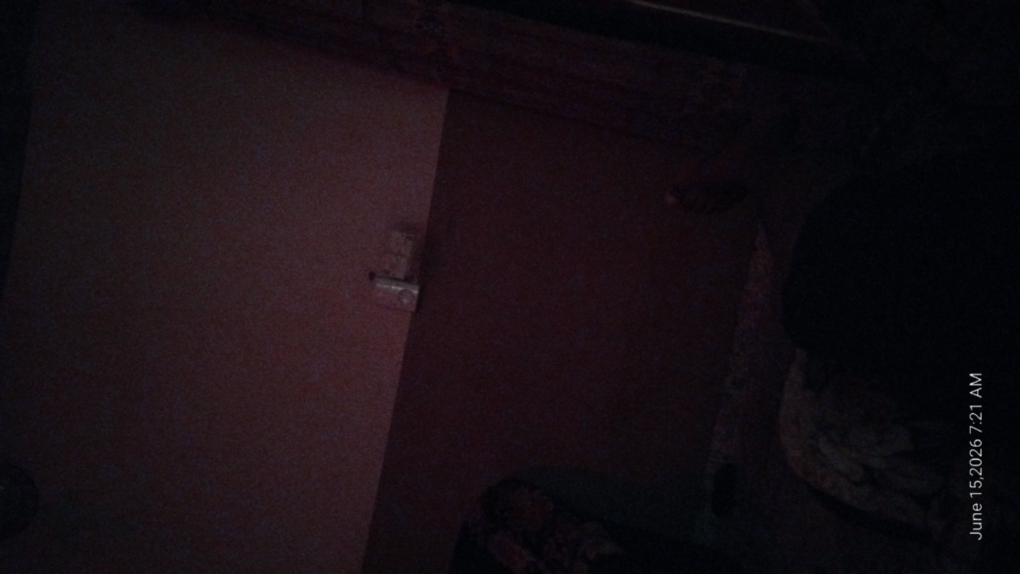 The image displays a dimly lit interior scene, possibly a bedroom, given the presence of a bed in the foreground, covered with a patterned fabric. The lighting is low, casting long shadows and obscuring finer details. In the upper portion of the image, on a horizontal surface, are several small, light-colored objects that appear to be containers or packages, possibly hygiene products or small electronic devices. The wall behind these objects has a faint, textured pattern. The overall color palette is dominated by dark reds and browns, with the lower section of the image being almost entirely black, suggesting the presence of a person or large, dark objects. The bottom left corner of the image contains a timestamp: "June 15, 2026 7:21 AM". The location of Howayi, Nigeria is mentioned, but there are no specific visual cues within the image to confirm this location.