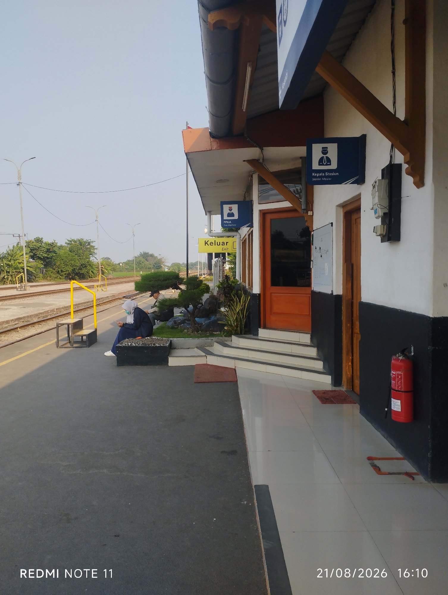 The image depicts a railway station platform in Karawang, Indonesia. The platform is paved and has train tracks to the left. A person wearing a headscarf and dark clothing is seated on a bench on the platform, looking at their phone. Another person is seated further down the platform. The station building has a white facade with black lower walls and a covered overhang supported by wooden beams. Signs on the building indicate "Kepala Stasiun" (Station Master) and "Keluar" (Exit). A red fire extinguisher is mounted on the wall near a wooden door. The date stamp on the image reads "21/08/2026 16:10". The sky is clear and blue, and there are trees and streetlights visible in the background.