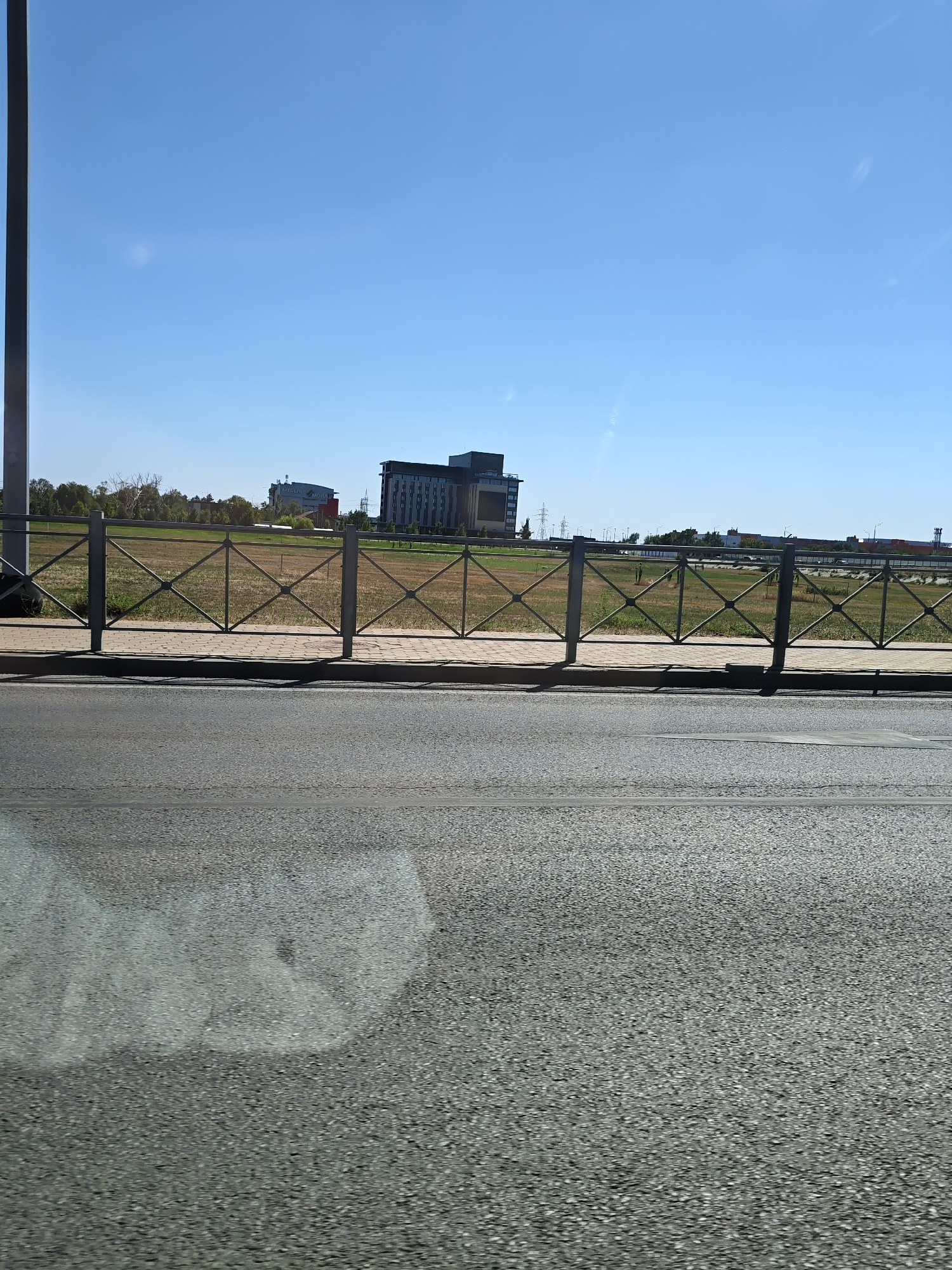 An exterior view of a modern cityscape under a clear blue sky. In the foreground, a grey asphalt road occupies the bottom half of the frame, with a light reflection on its surface. Behind the road, a metal fence with a diamond pattern separates it from a paved walkway. Beyond the walkway, a field of dry grass stretches out, dotted with scattered trees. In the background, several modern buildings are visible. On the left, a squat building with a red sign is situated amidst trees. To its right, a taller, multi-story building with dark facade and many windows stands prominently. Further to the right, another large, dark building with illuminated sections is visible. Electrical pylons and more buildings can be seen in the distance. The scene appears to be a developed area on the outskirts of a city, characterized by a mix of open space and modern architecture.