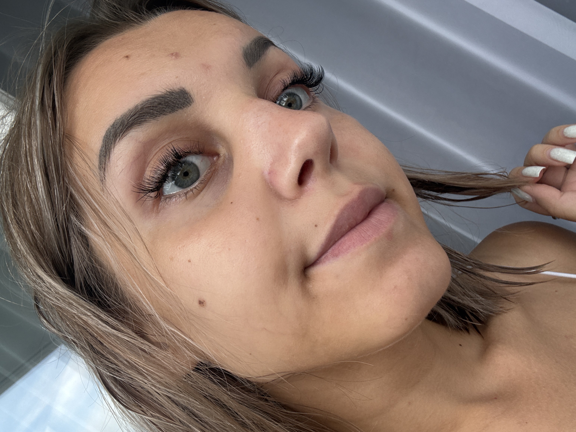 A close-up, angled shot shows the face of a young woman with light brown hair and light blue eyes. She is wearing artificial eyelashes and her eyebrows are dark and well-defined. Her skin is fair with a few freckles and beauty marks. Her lips are slightly parted and are a light pink color. She is touching her hair with her right hand, which has white nail polish on. The background is a gray, textured curtain. There is no indication of the city of Kholmsk, Russia, in the image.
