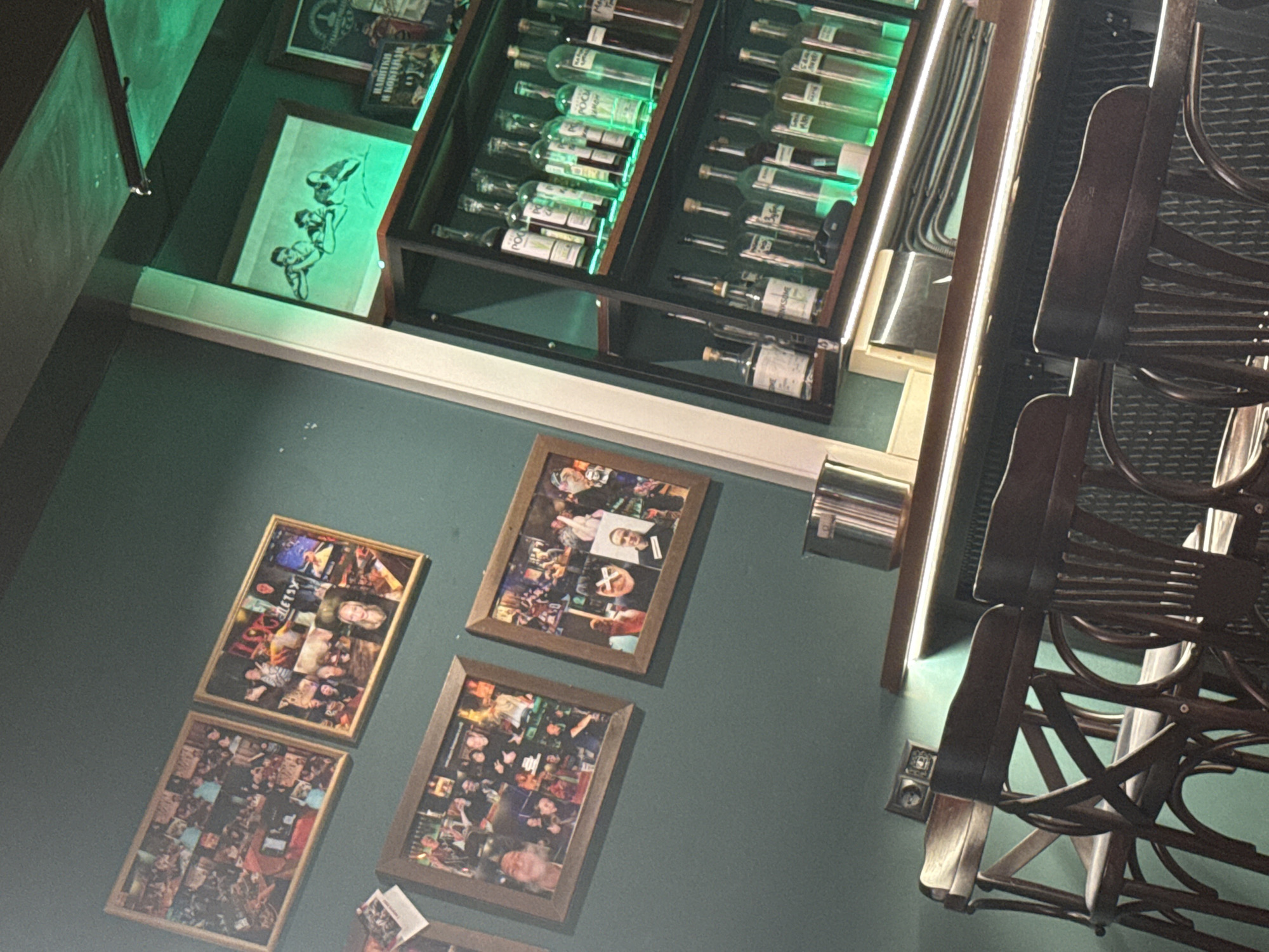 The image depicts an interior space with a green-tinted wall adorned with framed pictures of people. Above the pictures is a display case containing rows of bottled liquids, potentially alcohol, illuminated with green light. To the right of the display case, there are several dark-colored wooden chairs.
Given the details provided in the additional instructions, it's possible that the picture shows the interior of a restaurant in Moscow, Russia. However, it is not clear which one.