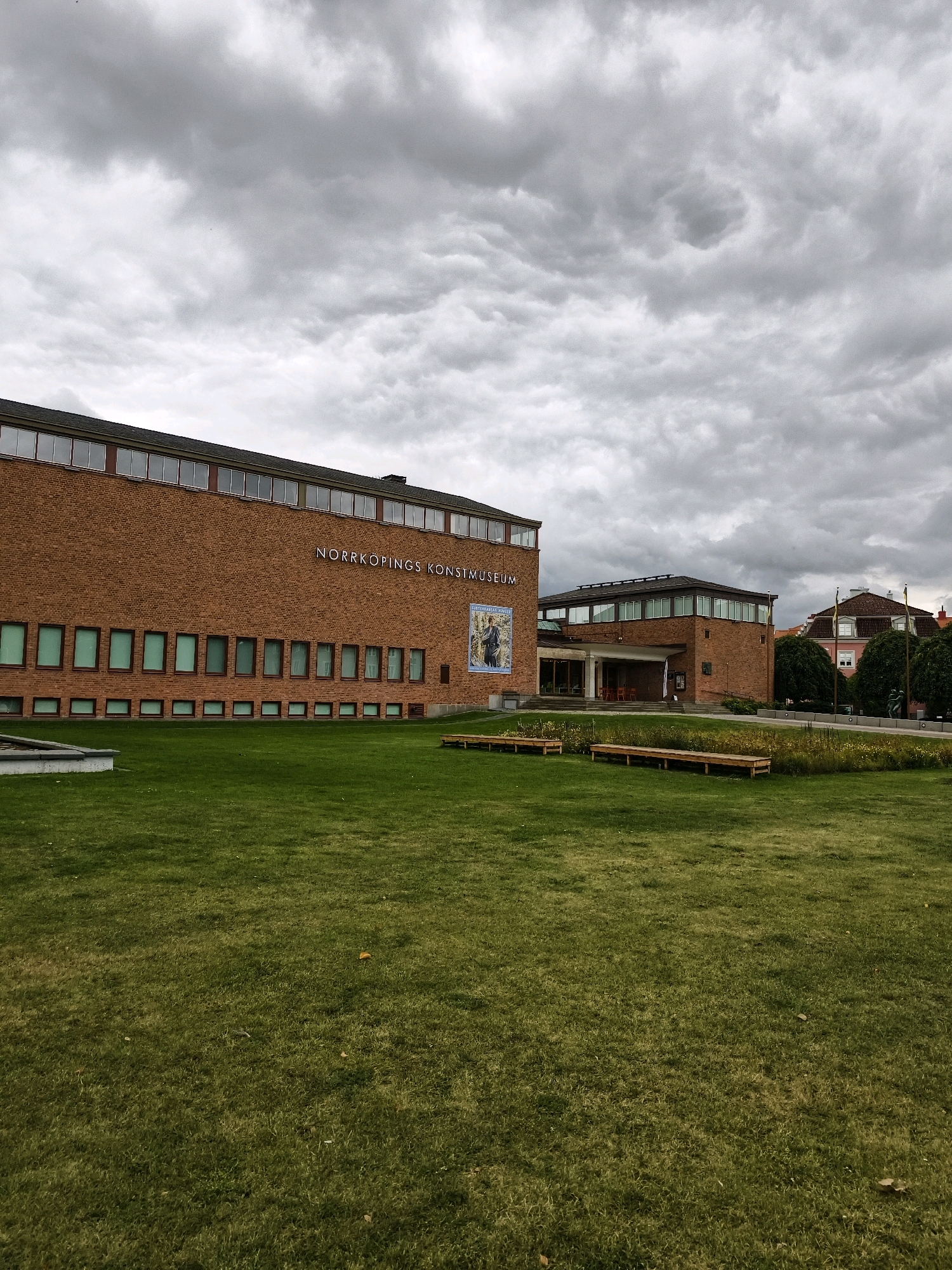 The image shows an exterior view of the Norrköping Art Museum, located in Norrköping, Sweden. The museum building is constructed of red brick and features a row of windows along its upper facade. A prominent sign on the building reads "NORRKÖPINGS KONSTMUSEUM". To the right of the main building, another section of the museum is visible, also made of brick. In front of the museum is a well-maintained green lawn, with several wooden benches placed on the grass. Beyond the museum and across a paved area with trees and some sculptures, residential buildings and more trees are visible. The sky is heavily overcast with dark, dramatic clouds, suggesting a possibly stormy or cloudy day. The scene appears to be devoid of any people.