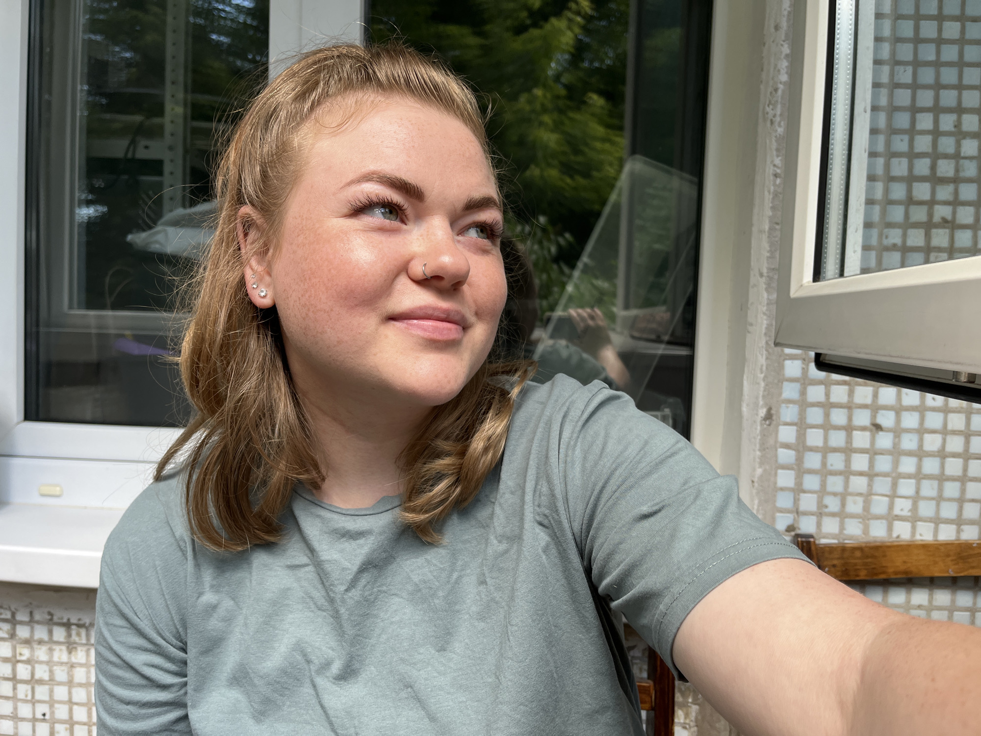 A young Caucasian woman with light brown hair pulled back and some strands framing her face is pictured. She is wearing a light green t-shirt and has multiple piercings in her left ear and a nose ring. The woman is looking to her right, with a slight smile on her face. She is seated outdoors, with a window and some greenery visible in the background. The wall behind her is tiled with small white squares. The scene appears to be in a residential area of Chaikovsky, Russia, given the architecture visible. The lighting suggests it is daytime, possibly late afternoon.