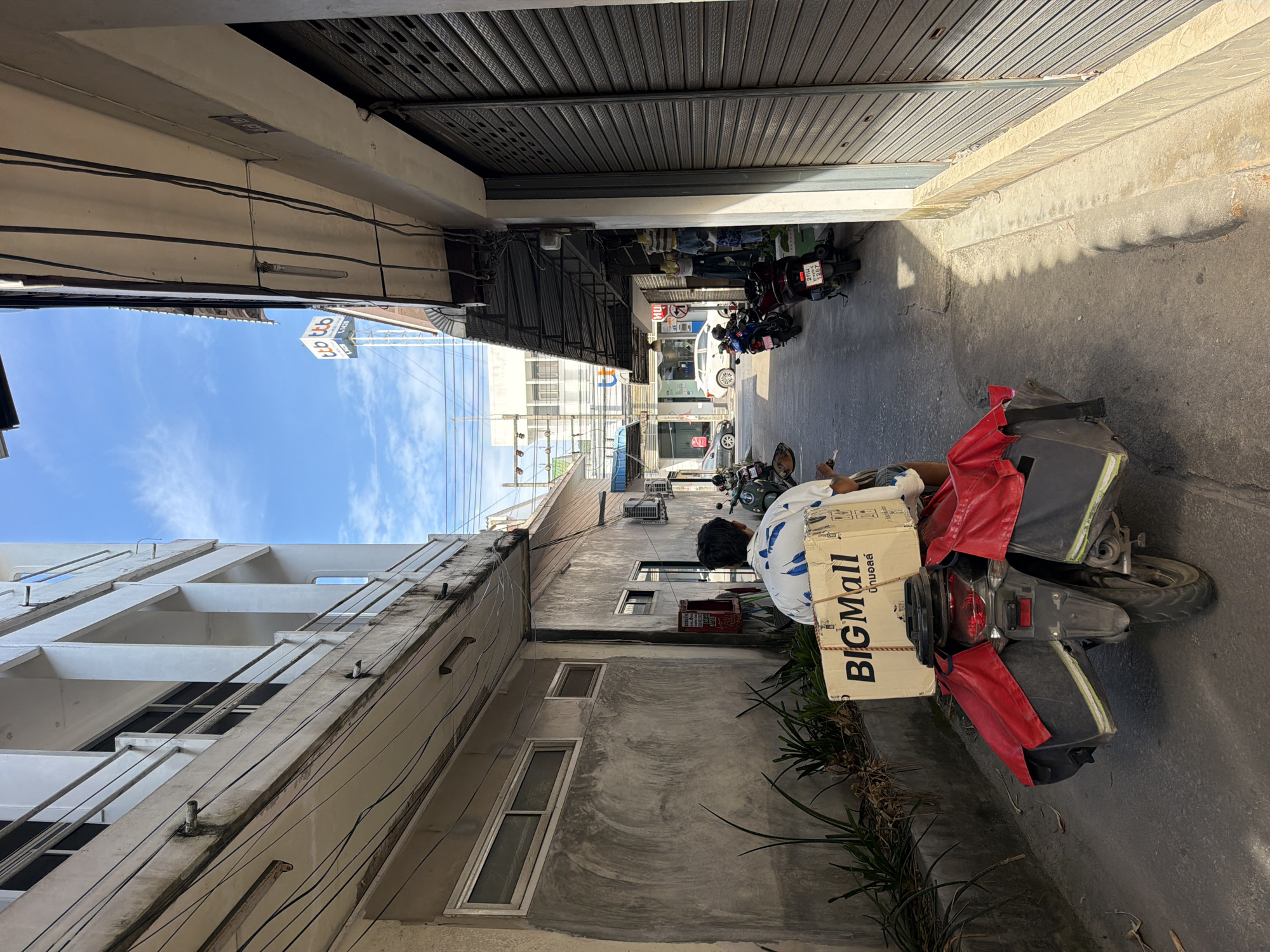 This is a street-level shot in the city of Ko Samui District, Thailand. A person riding a motorcycle is seen from behind, moving down a narrow alleyway. The motorcycle is equipped with large black panniers covered in red fabric. On the back of the motorcycle is a large cardboard box with "BIGMall" printed on it in black letters, and Thai script below. The alley is lined with buildings on both sides. The building on the left has a concrete facade and windows, with a row of plants growing along its base. The building on the right has a dark, corrugated metal shuttered storefront. Overhead, electrical wires are strung between the buildings. In the distance, a tall tower with "ttb" logo is visible against a blue sky with scattered clouds. Several other motorcycles are parked along the right side of the alley.
