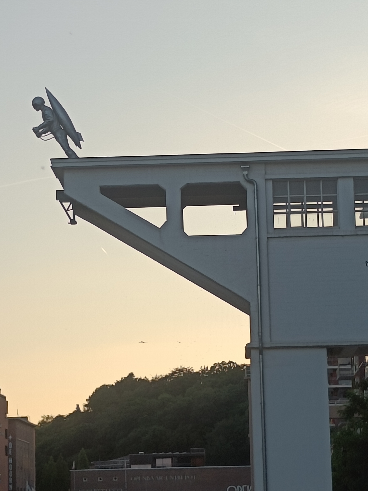 In Leuven, Belgium, a silver sculpture of a person in a jetpack is perched on the edge of a white building structure. The sculpture is facing left, with its left arm bent and holding onto the jetpack. The jetpack itself is shaped like a missile. The building features a large cantilevered section with an open rectangular window and a smaller square opening. Above the main structure, a row of windows with multiple panes is visible. In the background, a dense green forest covers a hilly landscape, with some residential buildings partially visible on the right side. The sky is a pale yellow, suggesting either dawn or dusk, with faint contrails from aircraft visible.