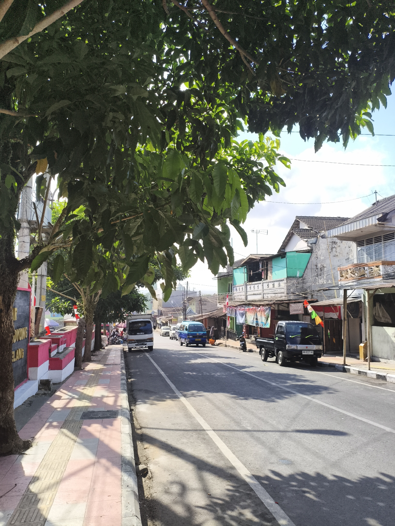 The image shows a street scene in Magelang, Indonesia. A sidewalk lined with trees runs along the left side of a paved road. On the sidewalk, there are several trees, a concrete barrier painted red and white, and a street sign. The road has two lanes of traffic and is bordered by buildings on the right. Several vehicles are on the road, including a white truck, a black pickup truck, a blue van, and other cars. The buildings appear to be a mix of residential and commercial establishments. The sky is clear with some clouds. The overall impression is a typical street scene in a moderately busy Indonesian town.