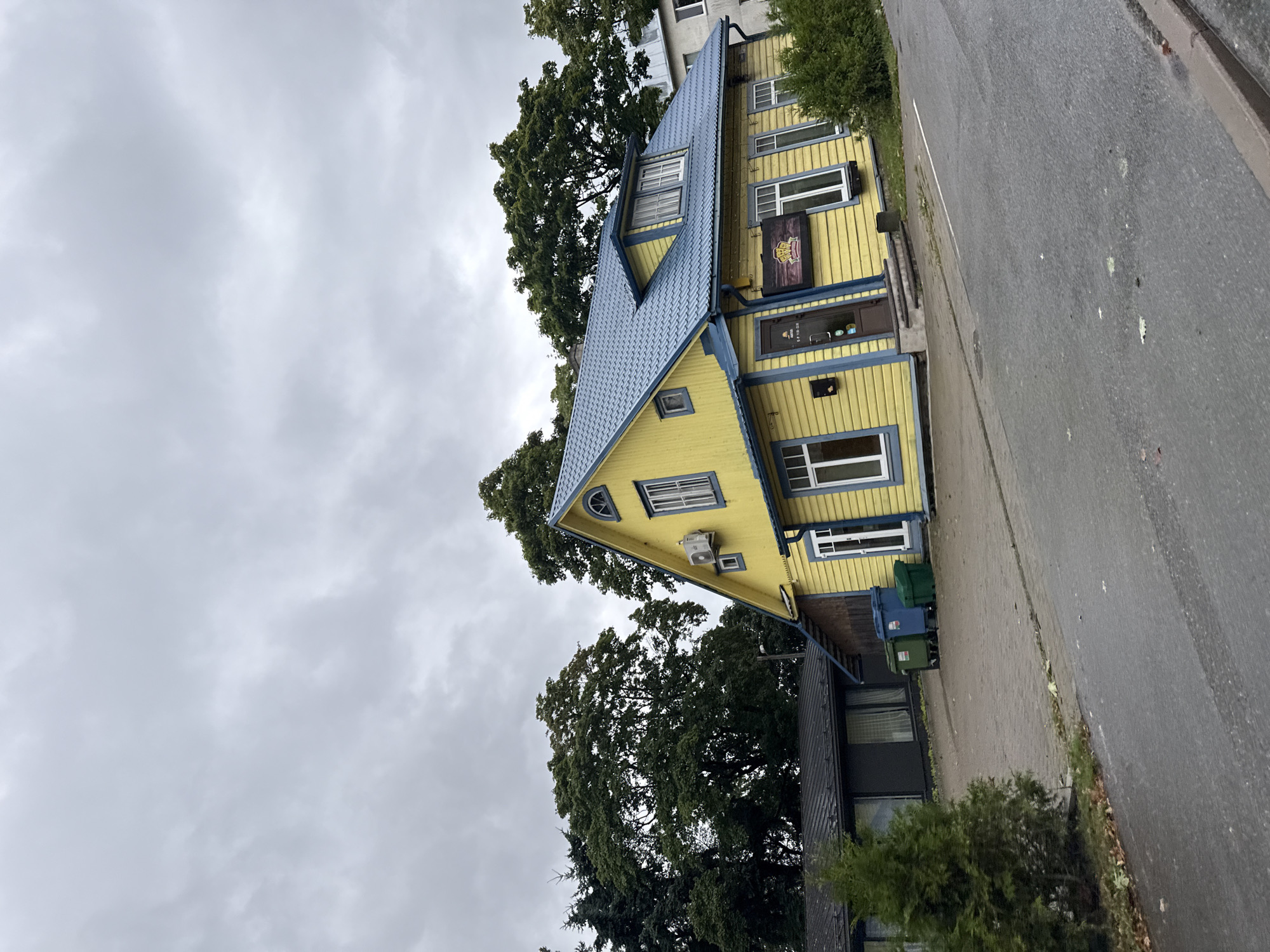 The image depicts a bright yellow, two-story house with a blue metal roof, situated on a paved area. The house has white-trimmed windows and a dark-colored door with a sign above it. To the left of the yellow house is a darker, single-story building with a metal roof. In front of the buildings, there are three trash bins, two of which are blue and one is green. A small evergreen tree is to the left of the trash bins. The ground in front of the buildings is paved with interlocking bricks. The scene is surrounded by lush green trees, and the sky is overcast with heavy gray clouds. The overall impression is that of a residential or possibly commercial property in a slightly damp, cool climate. The location is identified as Tallinn, Estonia.