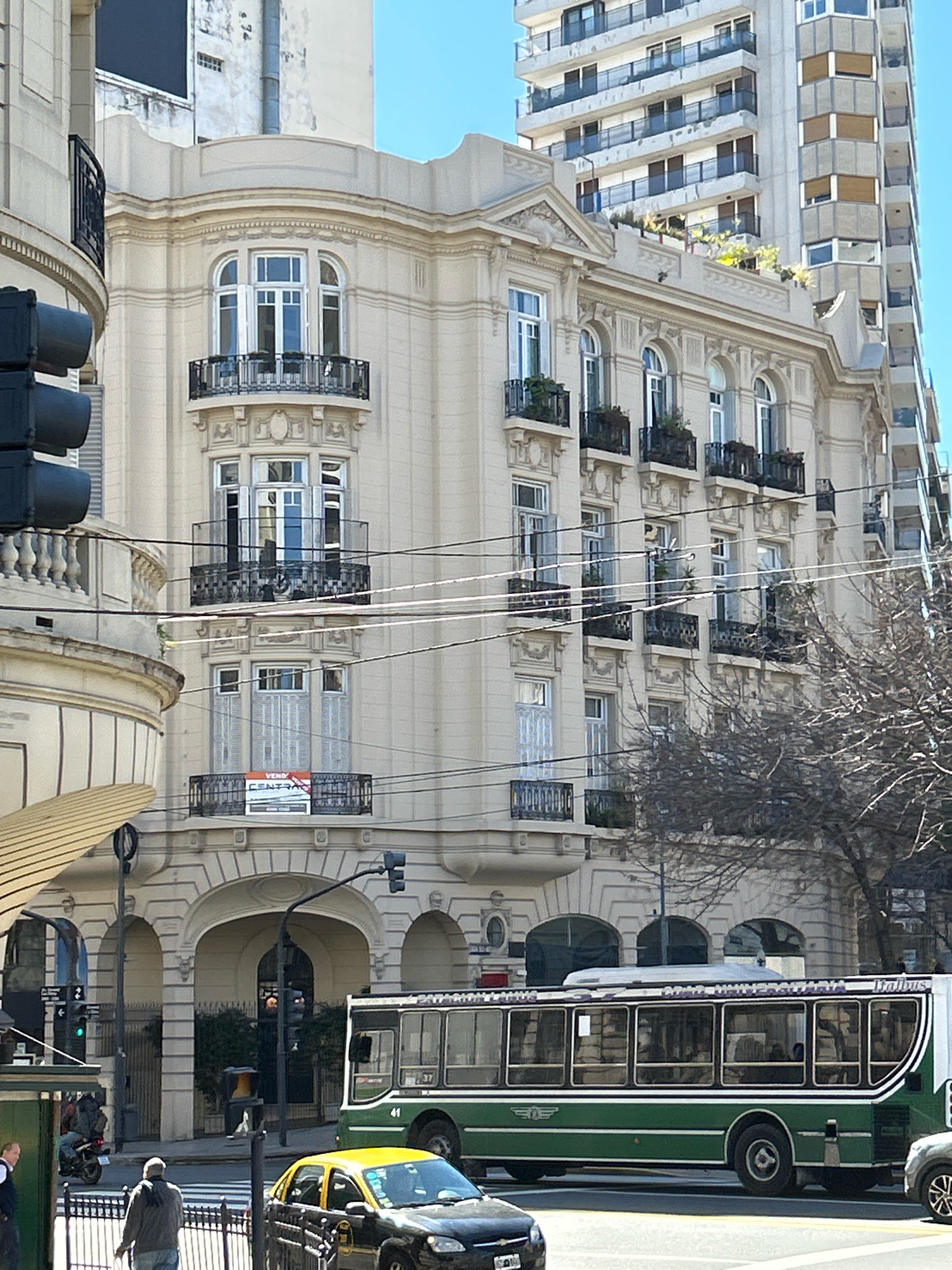 This is a street-level, eye-level, medium shot of a busy intersection in Buenos Aires, Argentina, likely on a sunny day given the clear blue sky and shadows. The foreground features a narrow sidewalk with a black metal fence. A man in a grey jacket and scarf is walking away from the camera on the sidewalk. To his right, a black and yellow taxi with its hazard lights on is stopped at a crosswalk. A green and white bus with the number "41" and "LINEAS ESTE" visible on its side is driving past the taxi. A light grey car is following the bus. Behind the bus, the street continues with more buildings and traffic signals.
The midground is dominated by a light beige, ornate building with multiple balconies featuring black metal railings. Some balconies have small potted plants. A sign that reads "VENDO" (for sale) is displayed on one of the balconies. Above this building, taller, modern apartment buildings with numerous windows and balconies are visible. Power lines crisscross the scene, casting faint lines across the buildings.
The background is a clear blue sky. The overall impression is of an active urban environment with a mix of historical and modern architecture. The location is identifiable as Buenos Aires due to the distinctive green and white buses (known as "Colectivos") which are a common mode of public transportation in the city, and the architectural style of the older buildings.