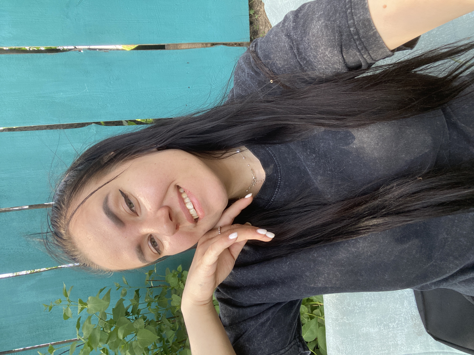 A young woman is posing for a selfie. She has dark hair and fair skin. She is wearing a dark-colored t-shirt. Her right hand is raised to her face. She is smiling. The background is a turquoise painted wooden fence and green foliage. The location is possibly in Chaikovsky, Russia.