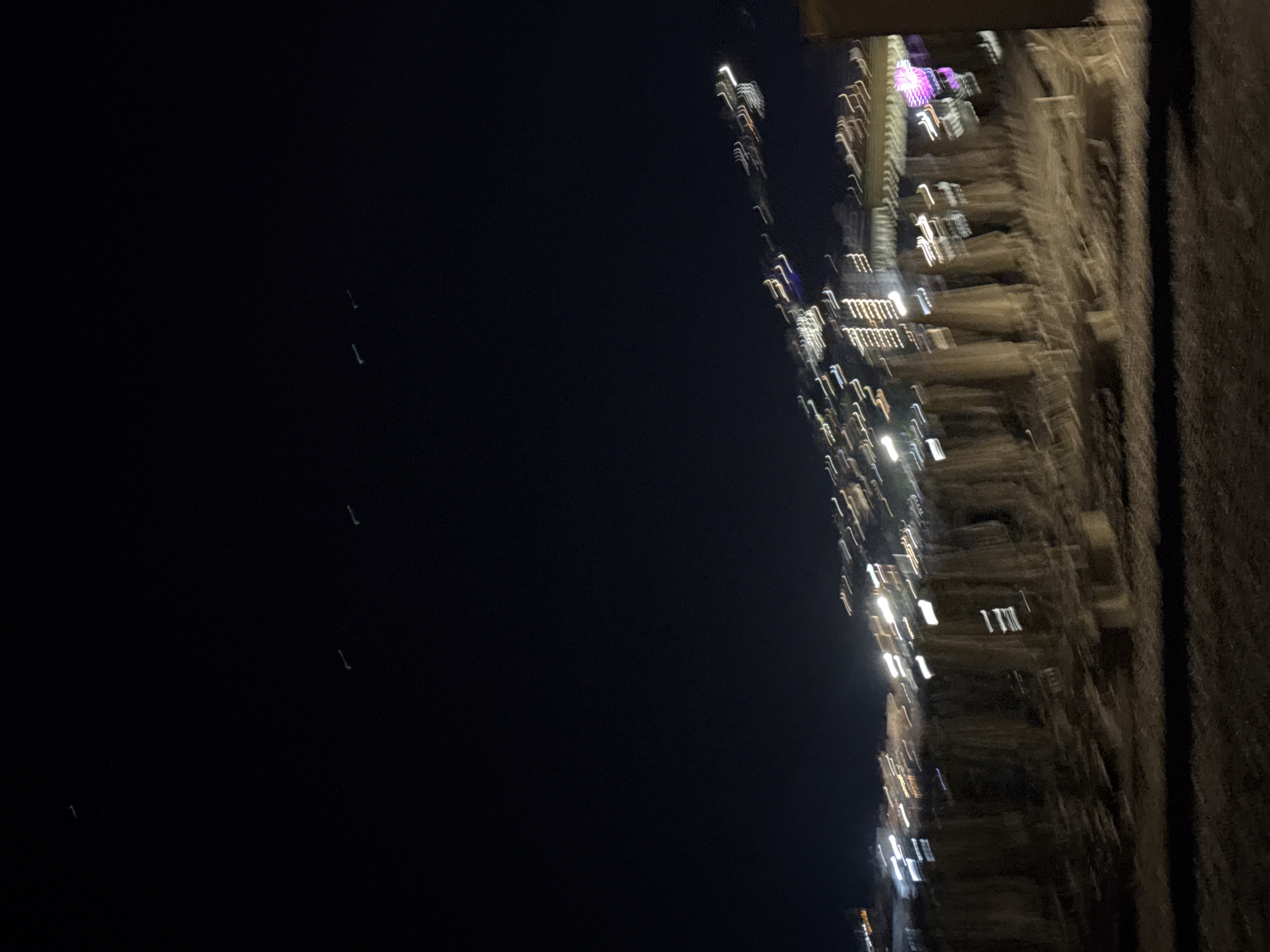 The image captures a blurry nighttime scene, likely of a beach or promenade area in Budva, Montenegro. The sky is a dark, deep blue, with several faint streaks of light that could be stars or distant aircraft. Below, a line of blurry structures, possibly beach umbrellas or temporary structures, are dimly visible, interspersed with streaks of light from buildings or streetlights. The lights are elongated and distorted due to motion blur, creating a dynamic, abstract effect. The ground in the foreground is dark and indistinct, suggesting sand or a similar textured surface. The overall impression is one of movement and a vibrant, albeit blurred, urban nightlife setting.