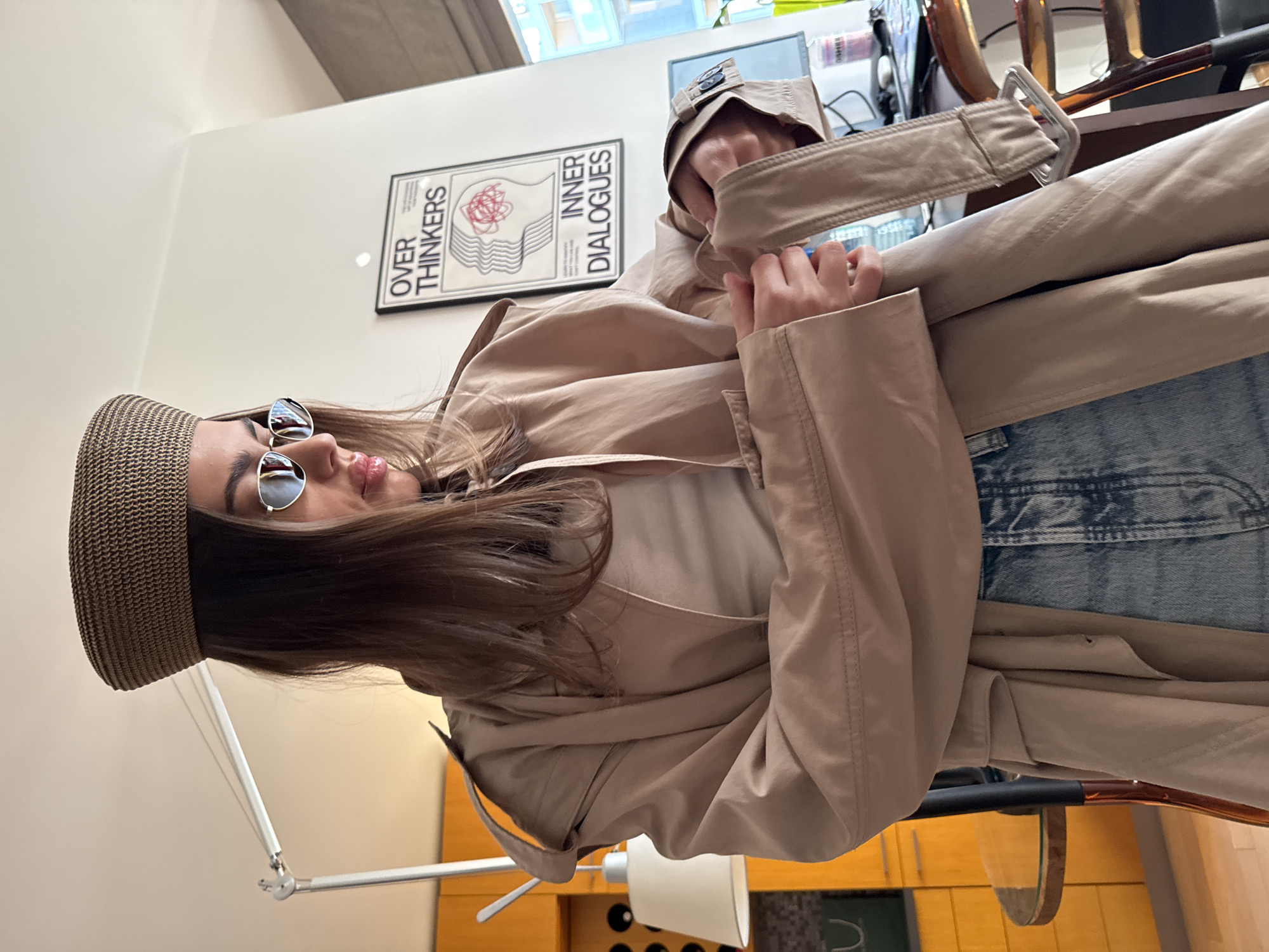 A medium shot depicts a woman wearing a straw hat, sunglasses, a white shirt, blue jeans, and a beige trench coat indoors. She is holding the trench coat's belt. Behind her on the wall is a poster with the words "OVER THINKERS" and "INNER DIALOGUES" printed on it. A standing lamp is visible in the foreground. The environment appears to be a modern living space or office. The location is not identifiable based on the image provided.