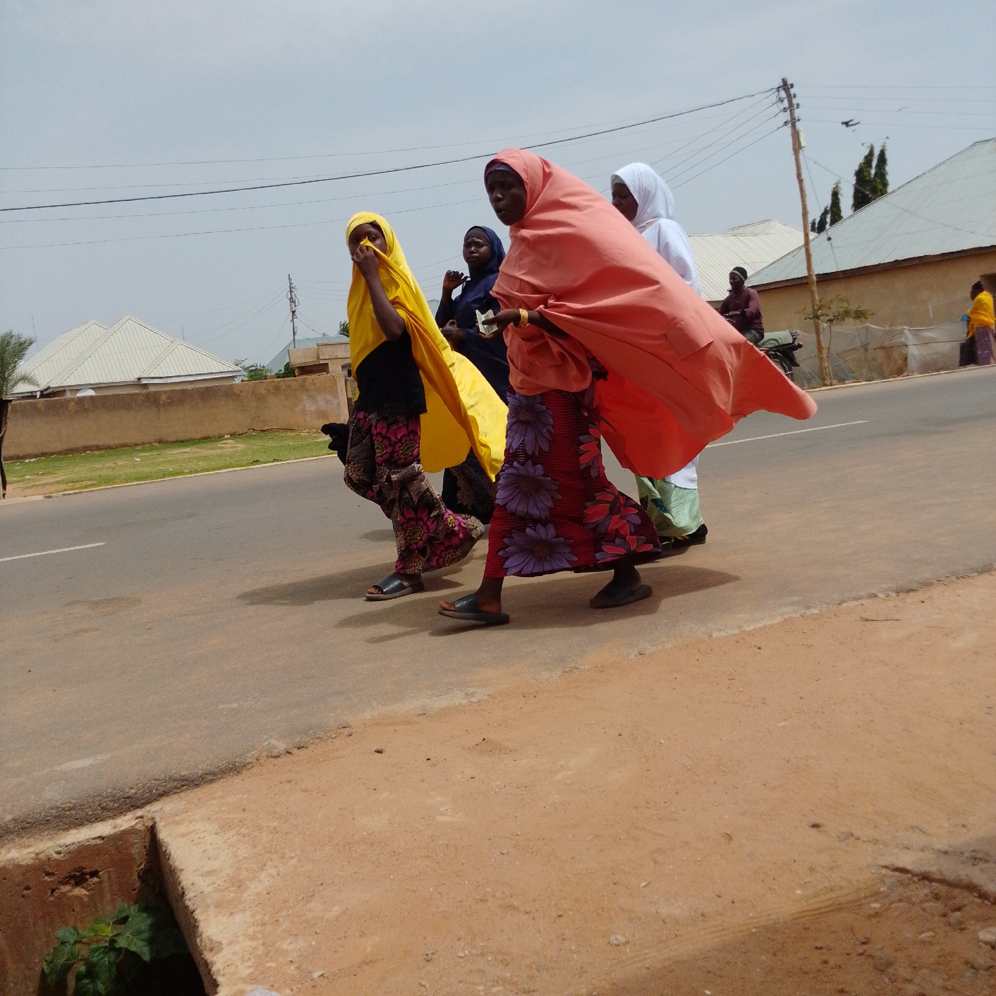 In Gombe, Nigeria, four individuals are walking on a paved road. The first person from the left is wearing a yellow hijab and a black shirt with a patterned skirt. Next to them, a person in a dark blue hijab is partially visible. The third individual is wearing a vibrant orange hijab and a floral-patterned skirt, holding something in their hand. The fourth person is in a white hijab and green clothing. In the background, buildings with light-colored roofs are visible. Utility poles with multiple wires stand along the side of the road. The sky is overcast, with a pale, hazy appearance. The ground is a mix of asphalt and dirt, with some patches of dry grass.