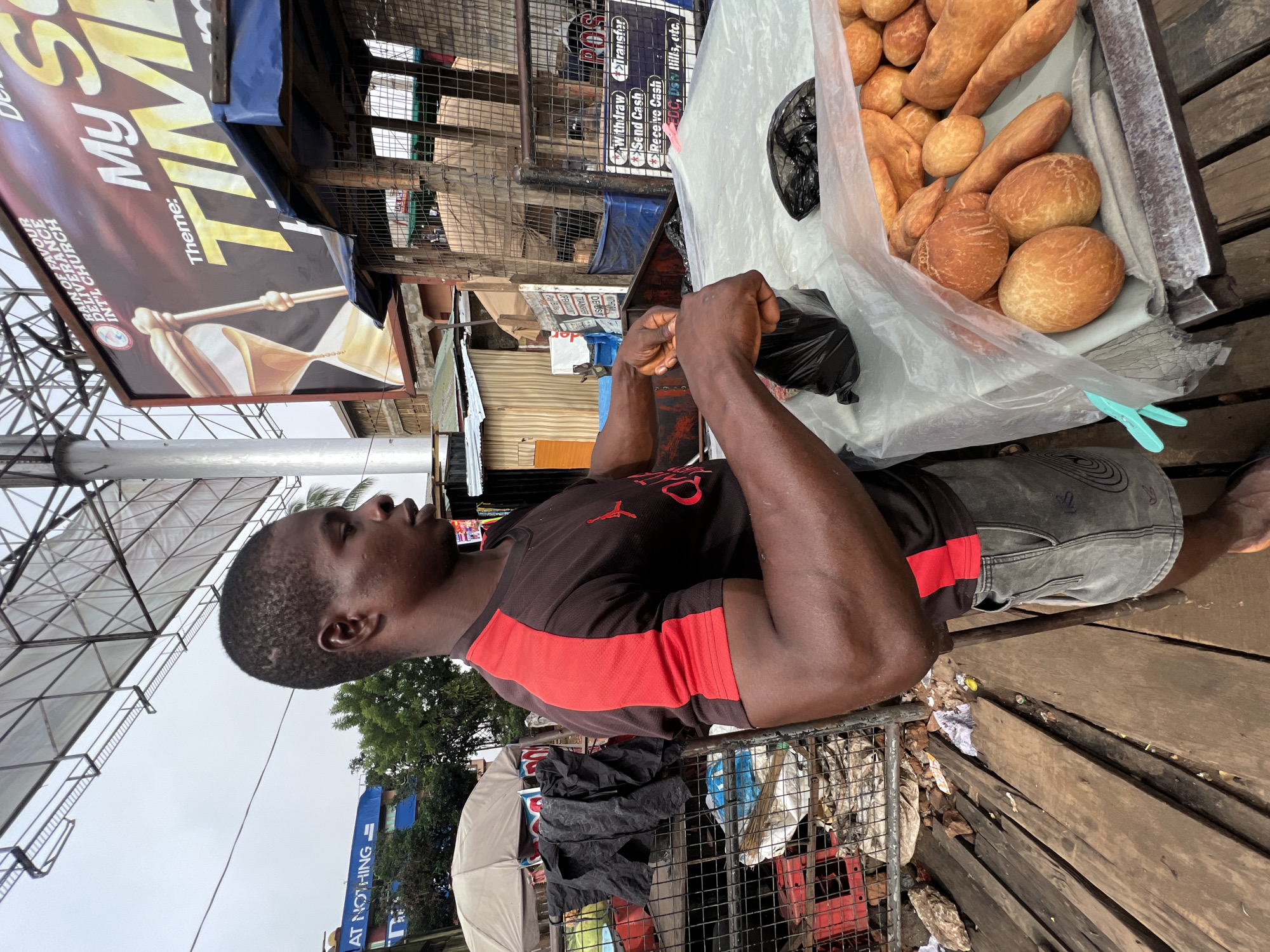 A man stands behind a table at an outdoor market in Nkpor, Nigeria. He is wearing a dark t-shirt with red accents on the sleeves and dark denim shorts. His arms are muscular, and he is holding a black plastic bag in his hands, appearing to be packaging something. In front of him, on the table covered with a clear plastic sheet, are numerous pieces of fried dough, varying in size and shape, some round and others elongated like small loaves. To the left of the man, there is a metal grate structure with black bags and red crates. In the background, a large billboard with the text "RAIN OF FAVOUR DELIVERANCE INT'L CHURCH" and "My TIME" is visible, as well as various market stalls and buildings. The ground beneath the table is made of weathered wooden planks, with debris scattered around. The sky is overcast.